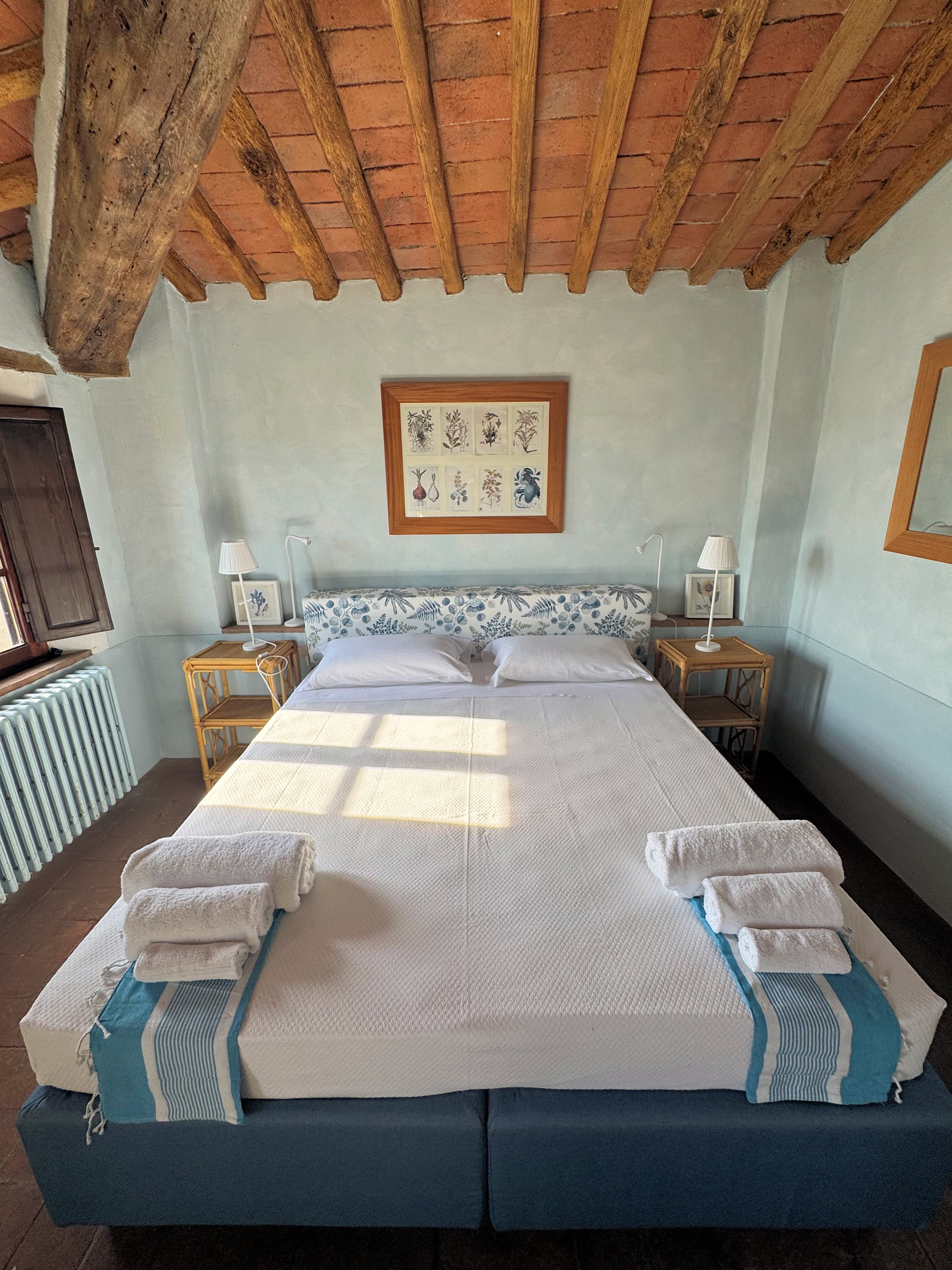 Rustic-chic master bedroom in a Tuscan farmhouse featuring exposed wooden beam ceilings, terracotta tiles, and a neatly made bed with folded towels.