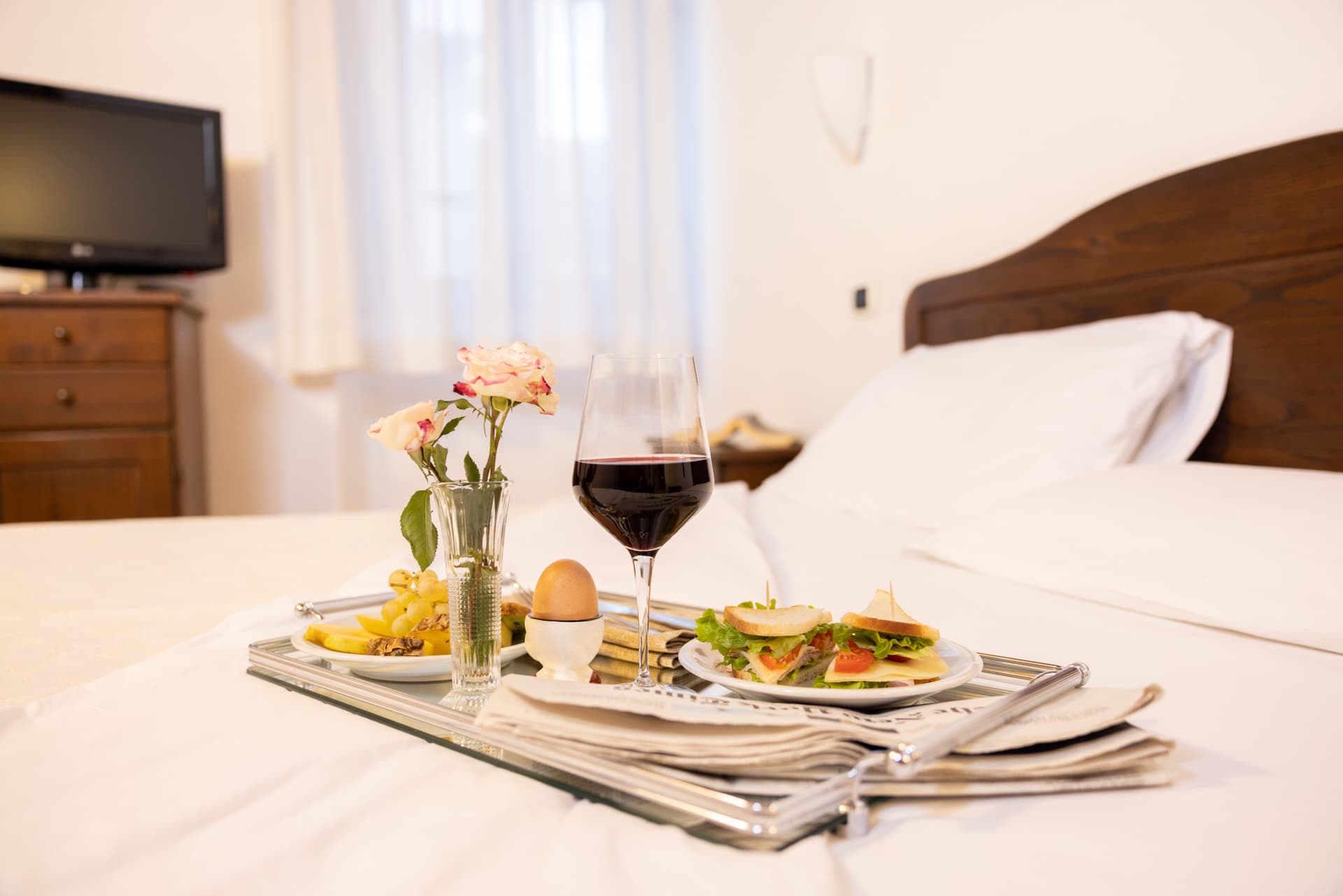 A silver tray with breakfast or a snack served directly on the bed of a hotel room. On the tray, which rests on white sheets, there is a glass of red wine, two sandwiches with lettuce and tomato, a soft-boiled egg in its egg cup, a small plate with grapes, and a small crystal vase with two roses. A daily newspaper is placed underneath the plates. In the slightly blurred background, you can see the bed's dark wood headboard with pillows, a window with light-colored curtains, and a television set on a wooden chest of drawers.
