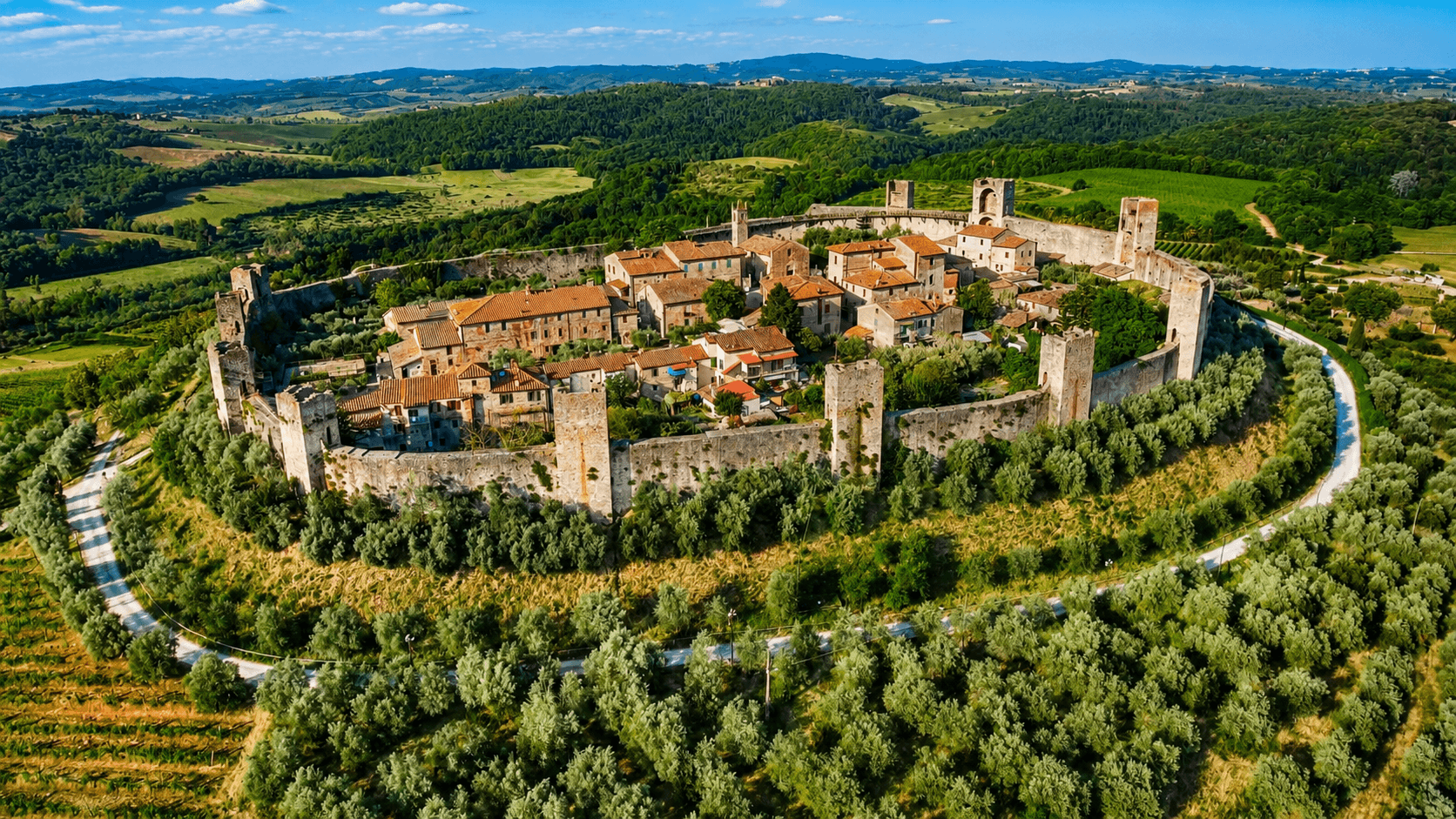 Aerial view of the medieval village of Monteriggioni with its circular walls and intact towers, surrounded by olive groves, vineyards, and green Tuscan hills