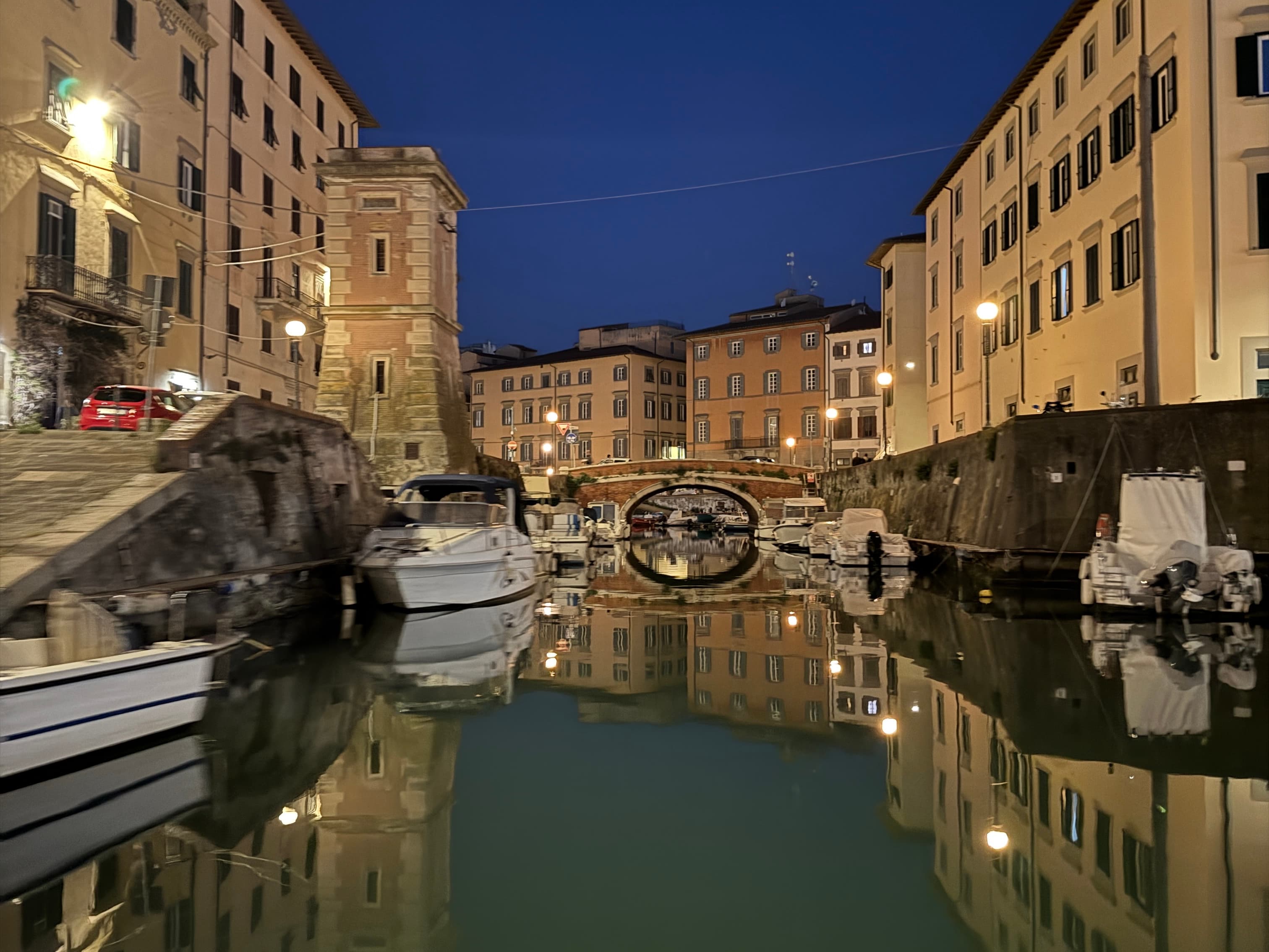 Livorno's Medici Canal at night with moored boats, illuminated brick arch bridge and historic buildings reflected in the still water