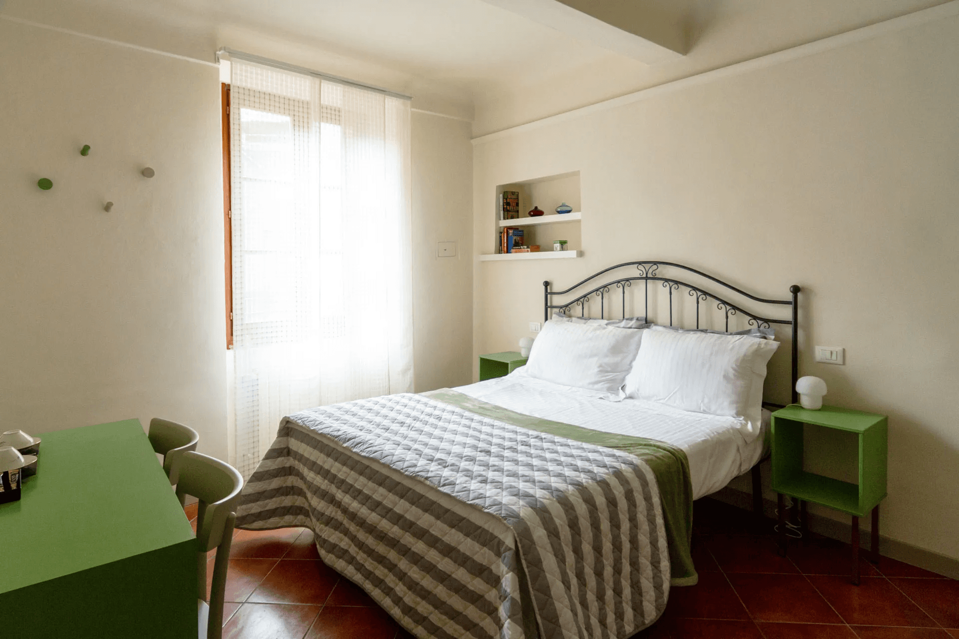 Camera da letto matrimoniale del B&B San Francesco a Siena, con letto in ferro battuto, copriletto a righe bianche e grigie, comodini verdi e pavimento in cotto toscano. Una grande finestra con tenda bianca illumina la stanza.