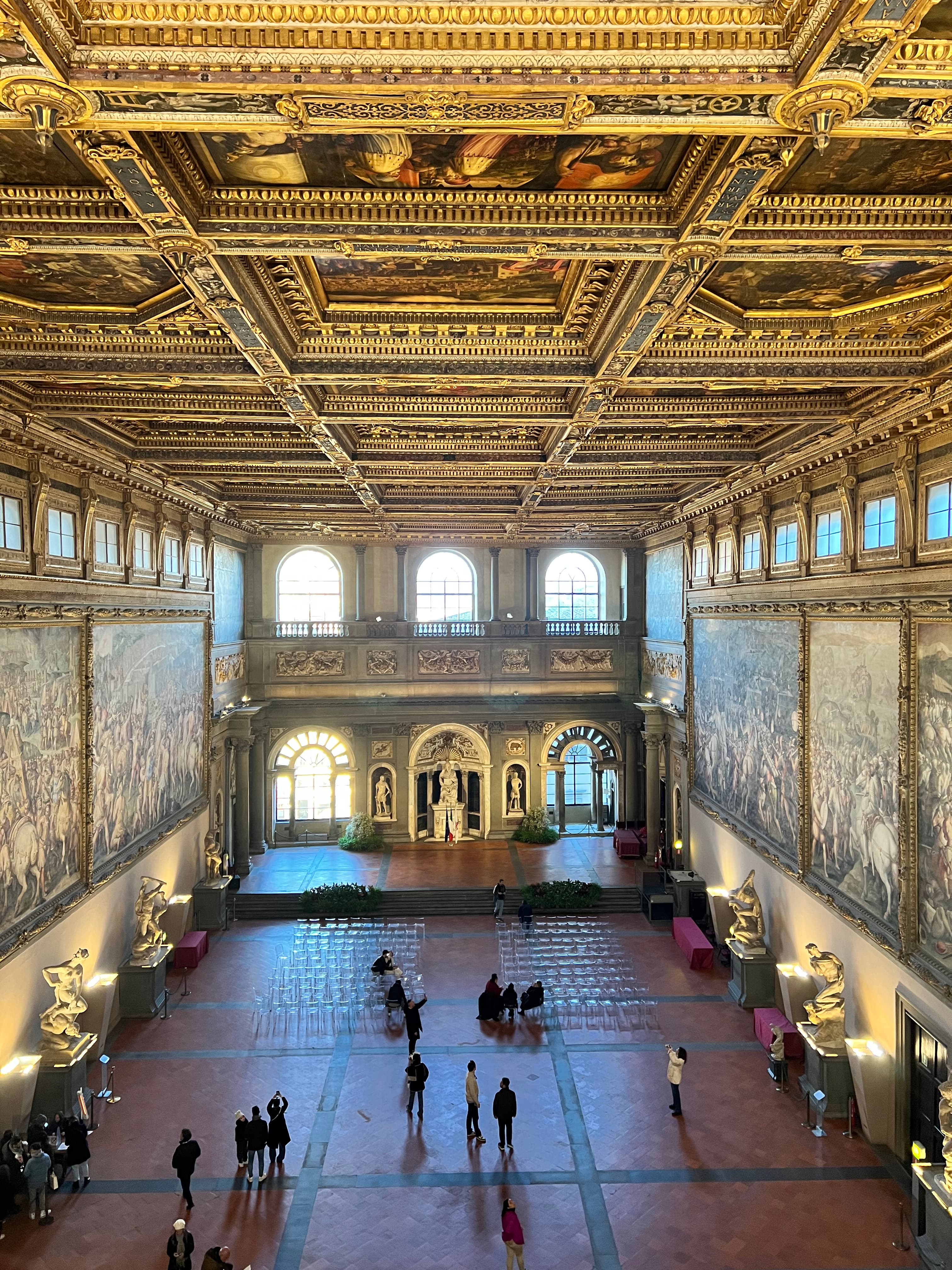Salone dei Cinquecento in Palazzo Vecchio con soffitto dorato a cassettoni, grandi affreschi rinascimentali e visitatori nella sala.