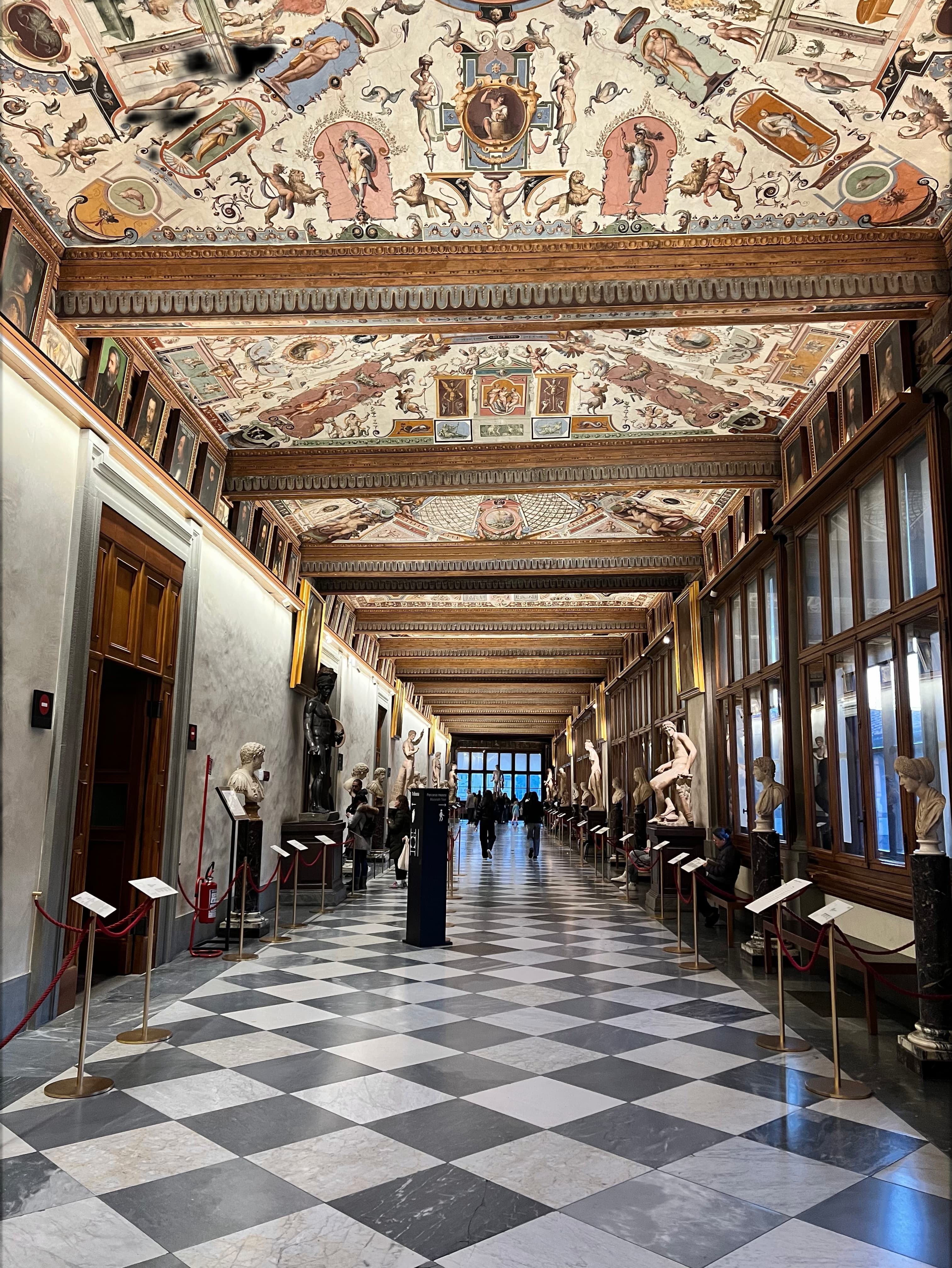Corridoio della Galleria degli Uffizi con soffitto affrescato rinascimentale, pavimento a scacchi in marmo e statue classiche lungo le pareti, visitatori sullo sfondo.