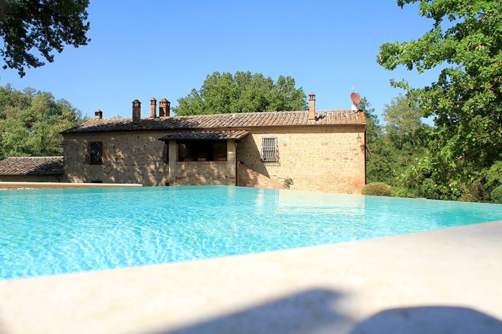 Foreground view of the clear, bright blue water of an outdoor swimming pool stretching towards the back facade of a historic stone farmhouse. The building features windows with iron grates, brick chimneys on the tiled roof, and a small covered loggia, surrounded by lush green trees under a clear summer sky.