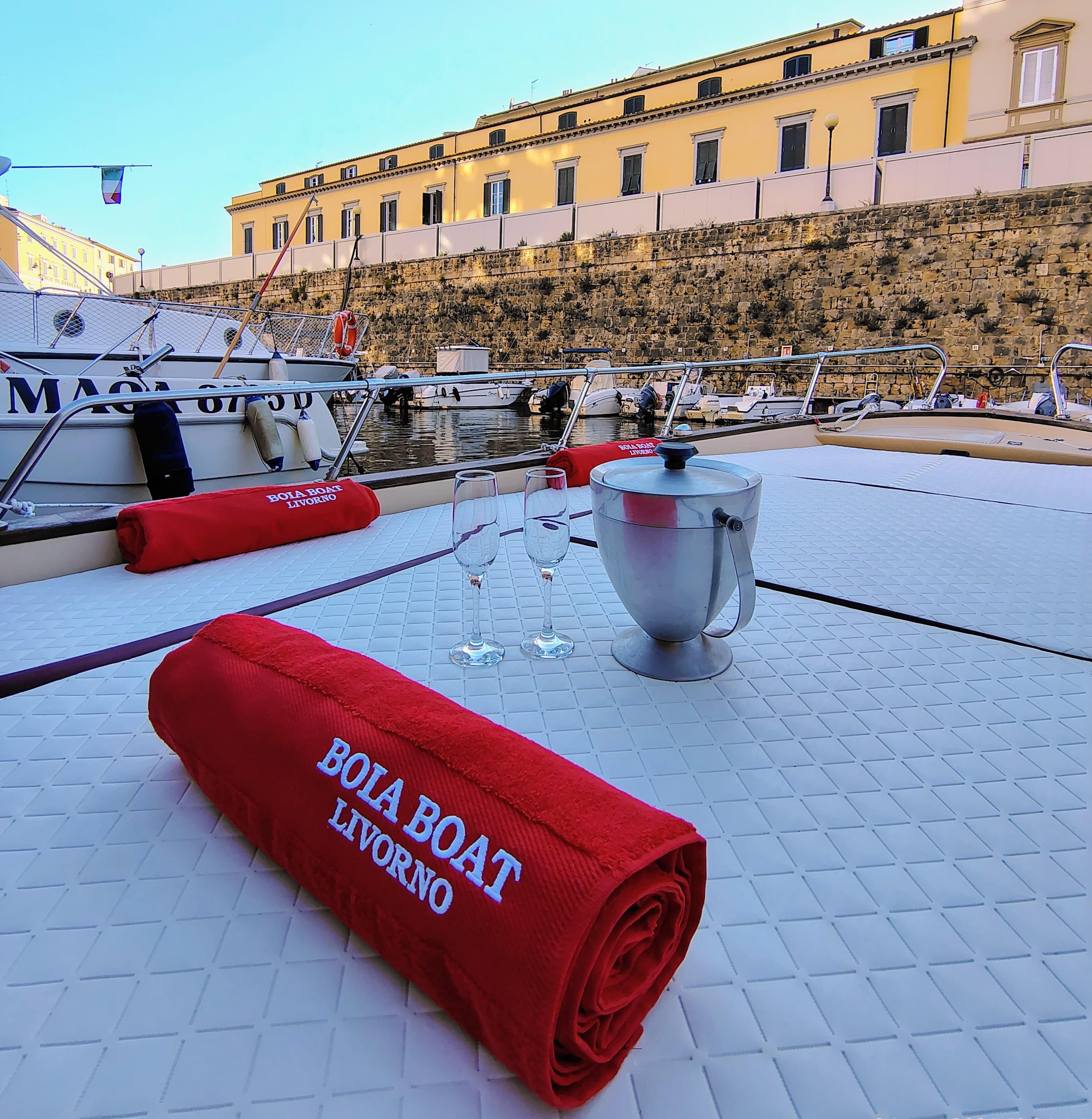 The bow of the Sorrento-based Boia Boat Livorno moored in the port of Livorno, with a rolled-up red towel with the Boia Boat logo in the foreground, two champagne glasses, and an ice bucket, with historic stone buildings in the background