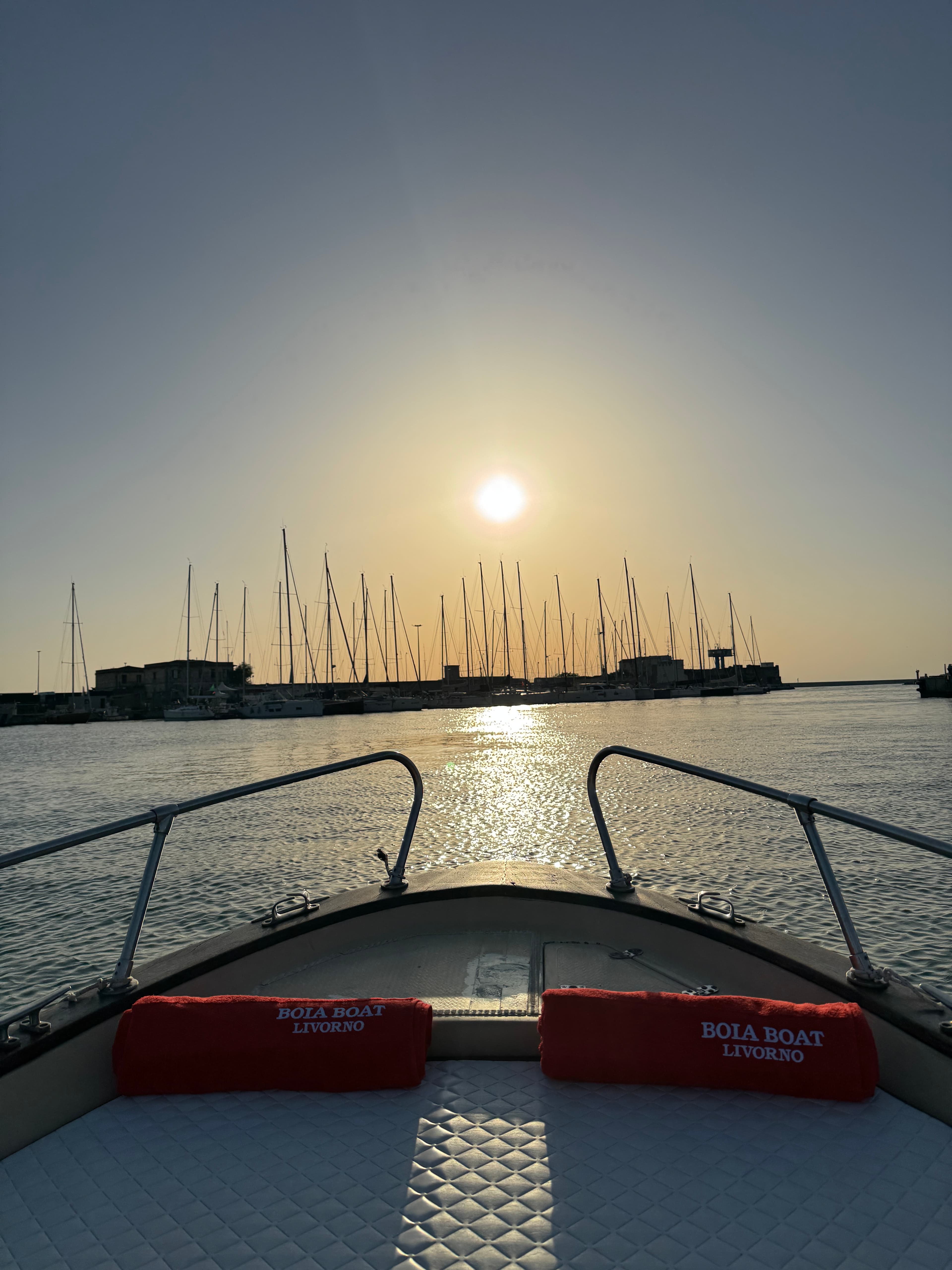 View from the bow of the Boia Boat Livorno toward the harbor with silhouettes of masts backlit at sunset, with two branded red towels resting on the metal railings of the sundeck