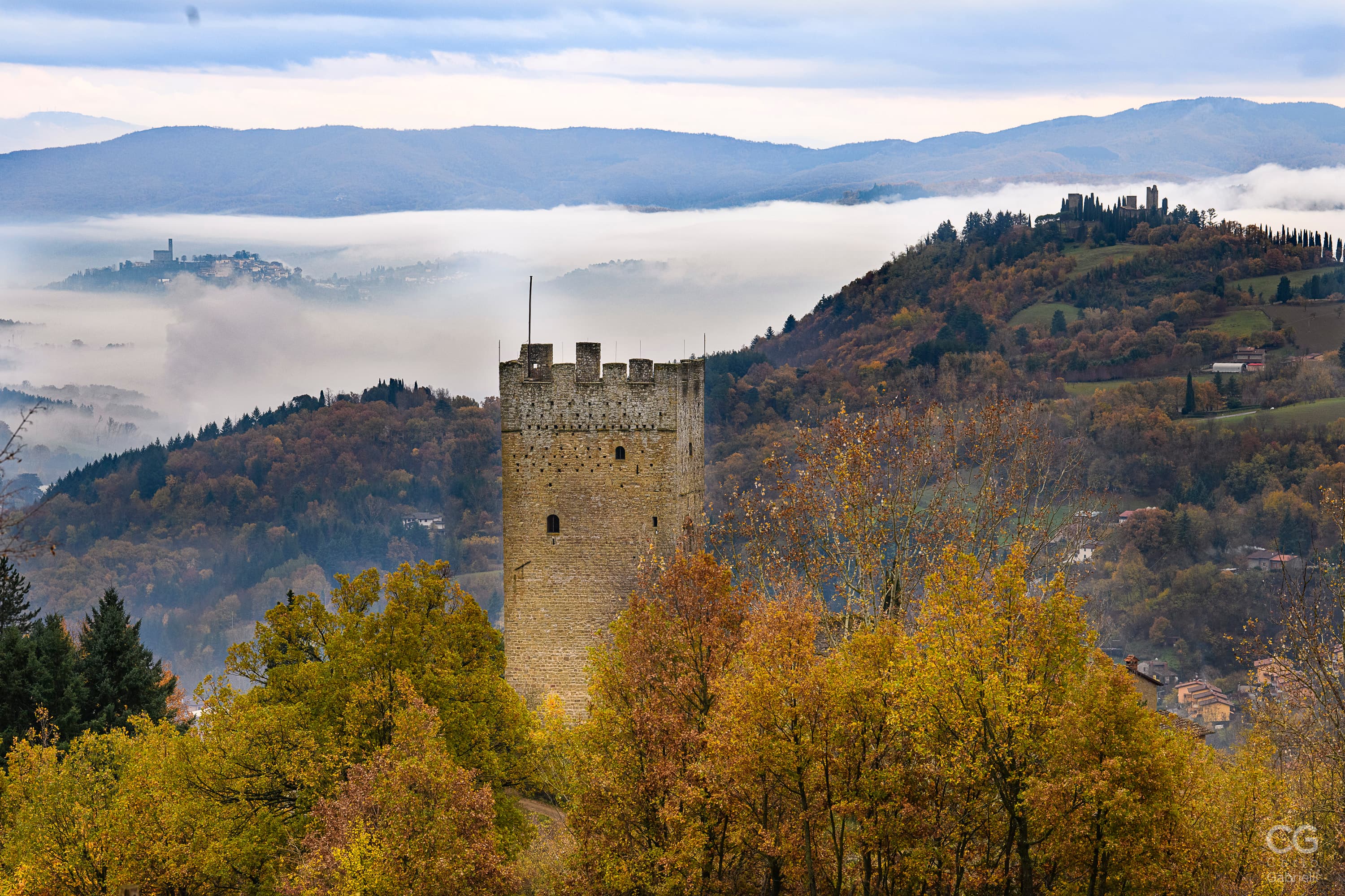 Medieval tower of the Casentino castles emerging from the autumn-colored trees, with a sea of ​​mist enveloping the Tuscan hills in the background