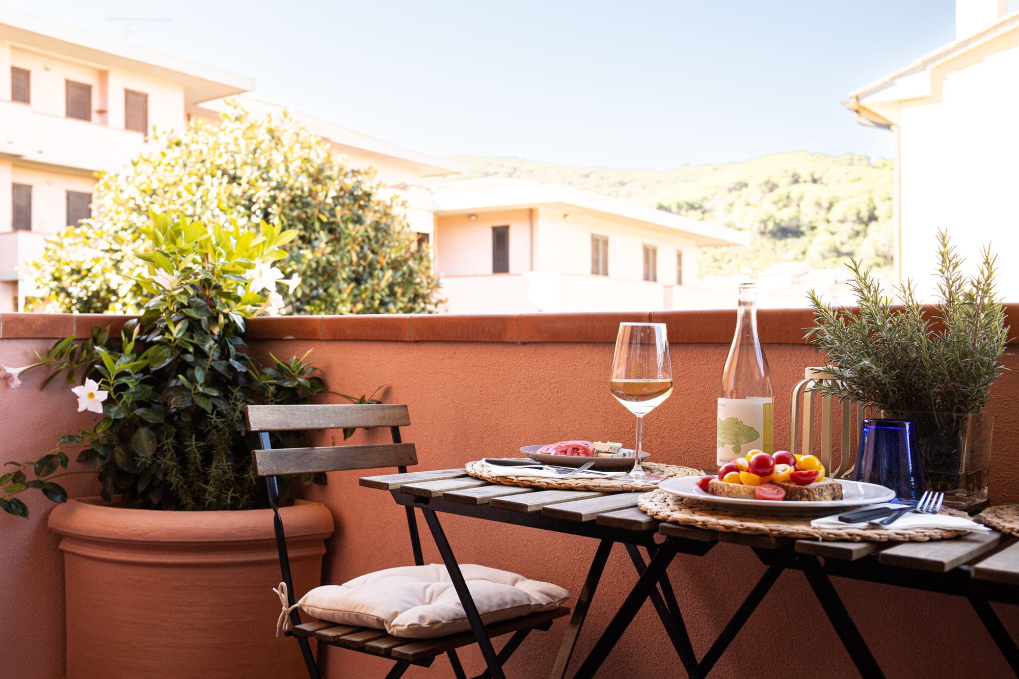 A wooden and wrought iron outdoor table is set on an apartment balcony for breakfast or an outdoor aperitivo. The table features plates with tomato bruschetta, a glass of white wine, a bottle, and a potted rosemary plant. Next to it stands a folding chair with a beige cushion and a large terracotta pot with flowering plants. In the background, buildings and green hills are visible under a clear sky.