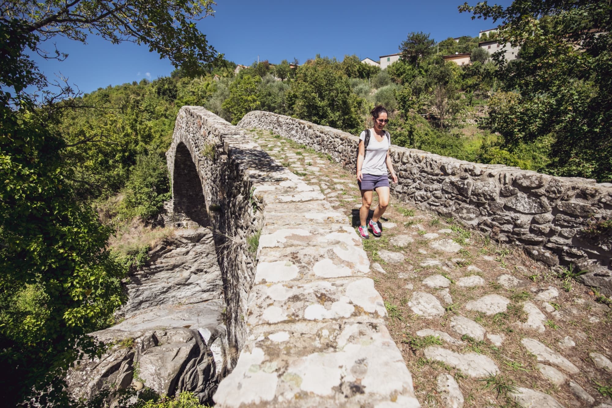 Escursionista attraversa il ponte romanico di Groppodalosio, immerso nel verde delle colline lunigianesi, lungo un antico tratto della Via Francigena.