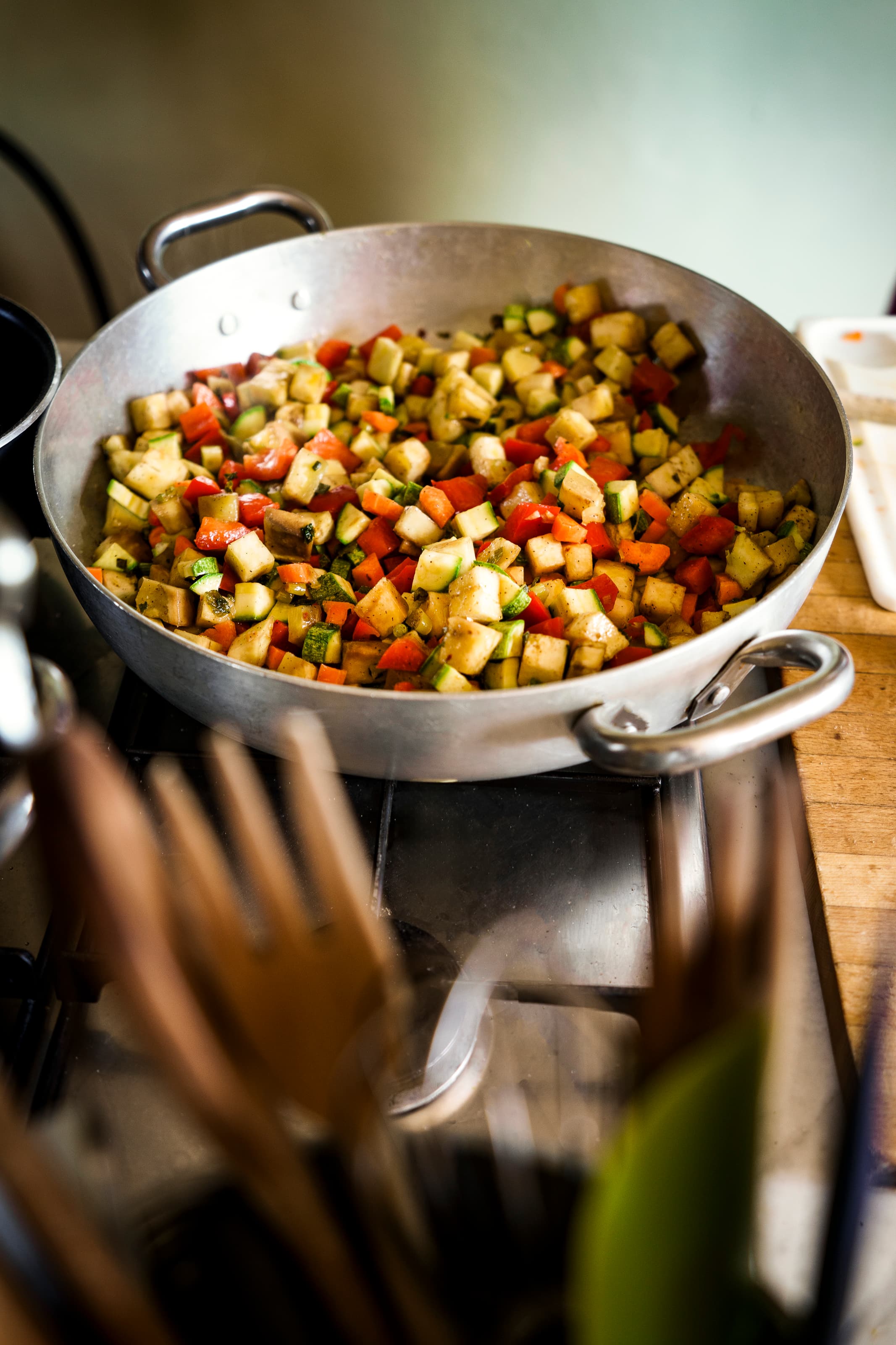 Padella professionale sul fuoco con verdure miste a cubetti tra cui zucchine, melanzane e peperoni rossi, durante una lezione di cucina toscana tipica