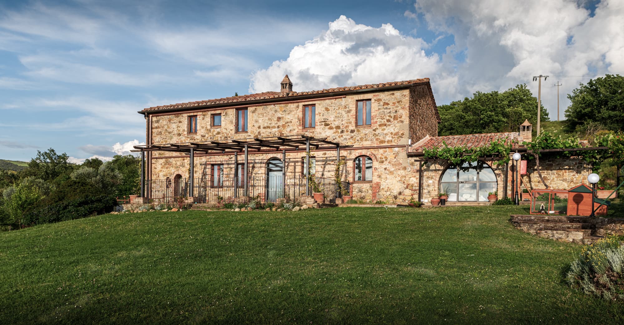 Facciata di un tipico casale in pietra toscano con ampio giardino a prato verde, pergolato in legno e cielo nuvoloso.
