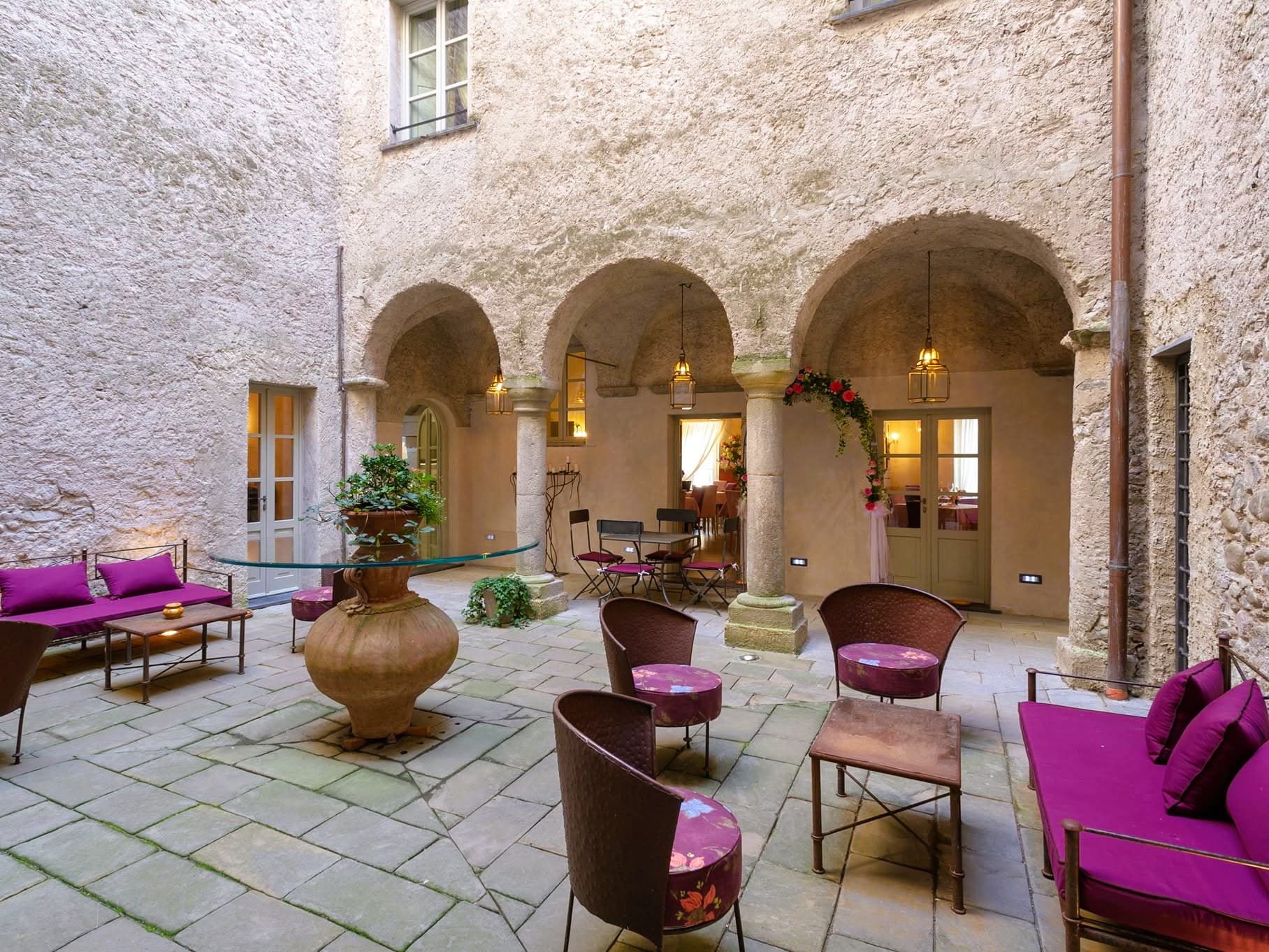 Cortile interno del Castello di Pontebosio con porticato ad archi in pietra, colonne medievali, lanterne accese e salottini con cuscini fucsia, sede delle visite guidate in Lunigiana