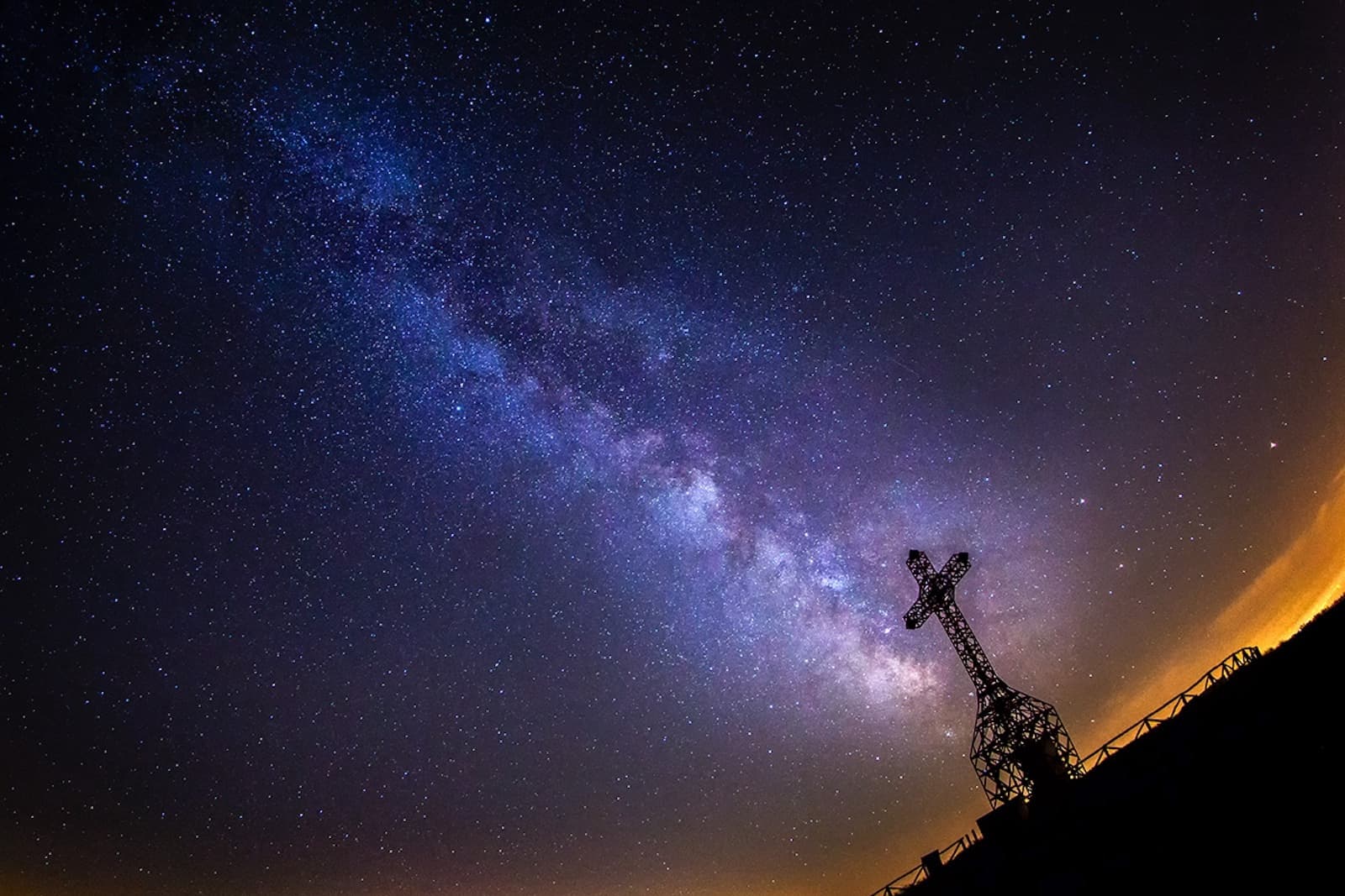 Silhouette of the metal Pratomagno Cross against the starry night sky with the Milky Way visible in shades of blue and purple, an orange glow on the horizon on the right side