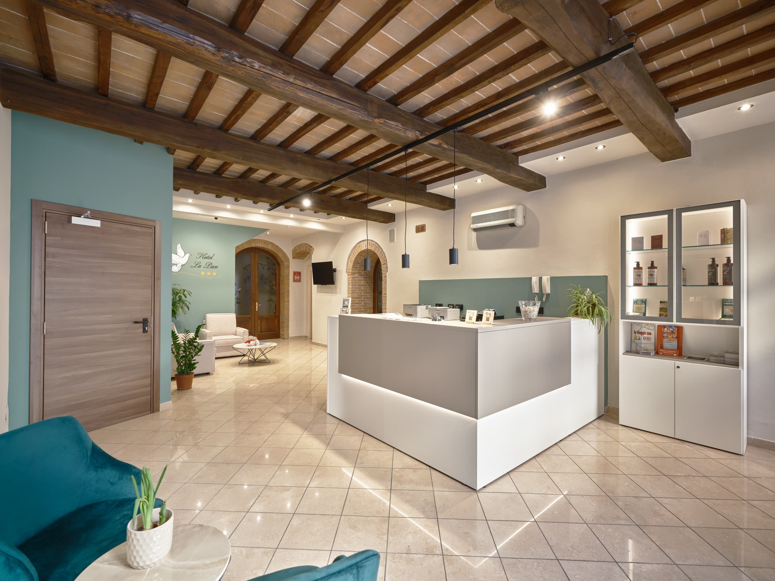 A modern and welcoming hotel reception that blends rustic elements, such as a ceiling with exposed wooden beams, with contemporary furnishings. The space features light-colored walls with sage green accents and a polished tiled floor. Dominating the center is a large, minimalist reception desk in white and dove gray, illuminated by LED lighting. To the right is a display cabinet showcasing products for sale, while to the left is a lounge area with velvet armchairs and a small table. The 'Hotel La Pace' logo can be seen in the background. The lighting is provided by modern spotlights and hanging pendant lamps
