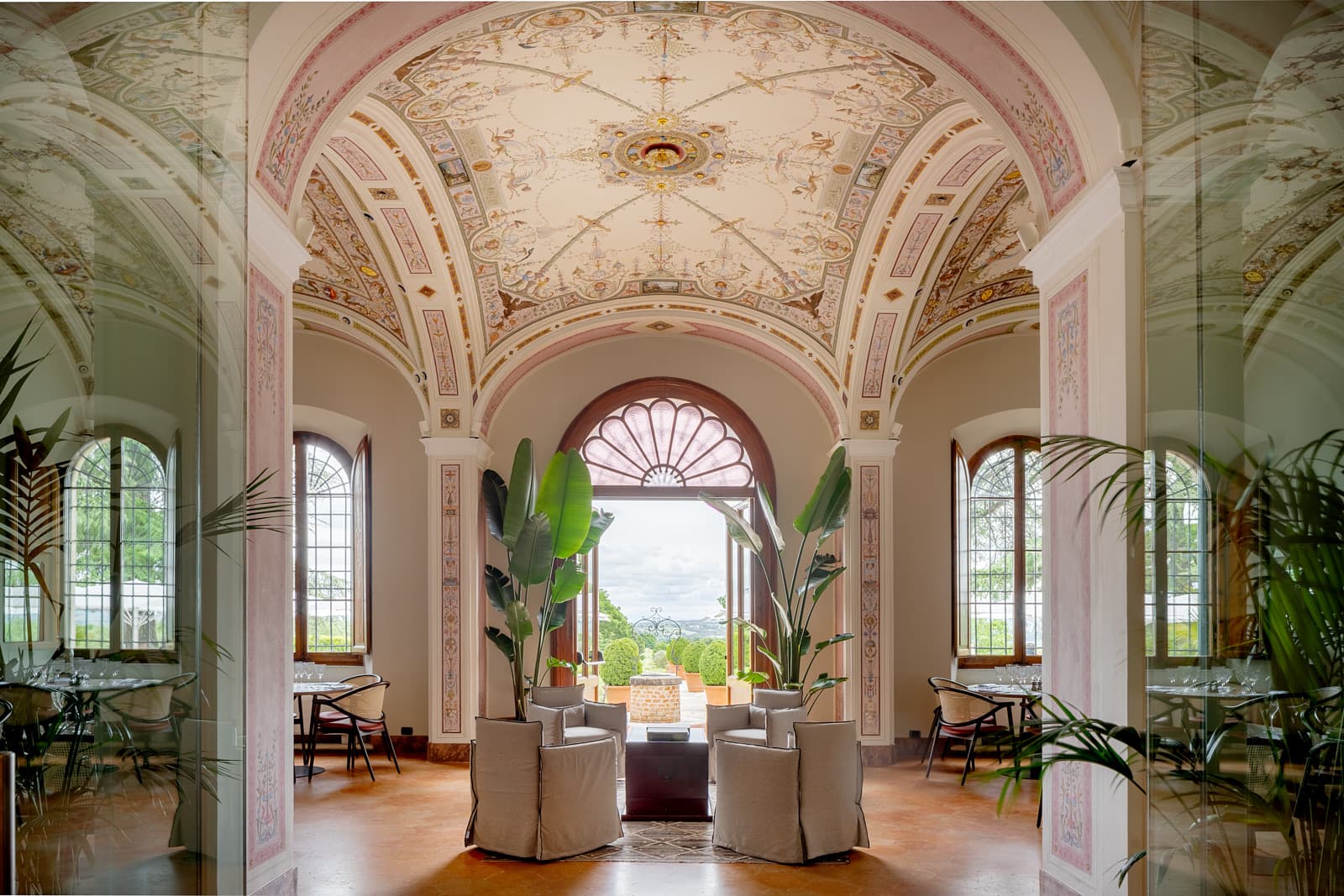 pacious and monumental grand hall of a historic Tuscan villa featuring magnificent barrel-vaulted ceilings completely adorned with period frescoes in soft pastel tones. At the center of the room, an elegant lounge area features four light gray fabric design armchairs arranged around a central coffee table, flanked by large potted Strelitzia plants. In the background, a large arched doorway with a sunburst-patterned lunette opens to the outside, offering a view of manicured gardens and the surrounding countryside. On either side, large arched windows illuminate the space, showcasing elegantly set dining tables. The floor is made of traditional terracotta, and large glass partitions in the foreground reflect the indoor greenery.