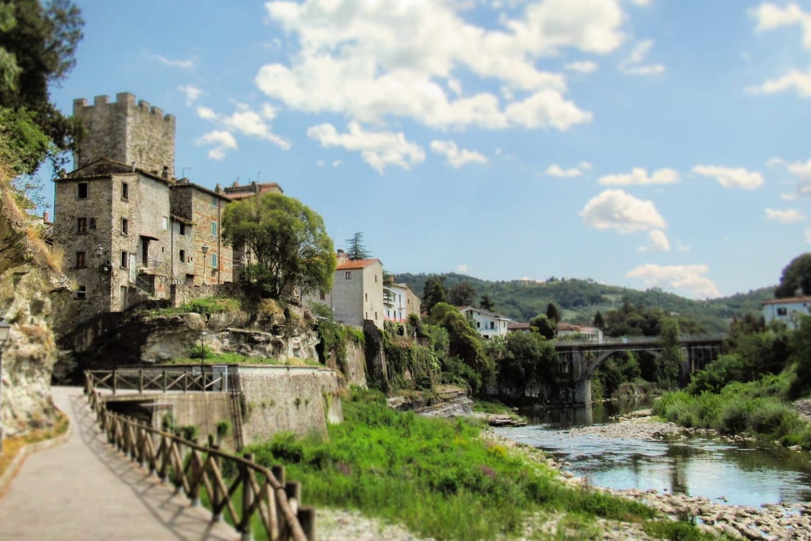 Torre medievale del Castello di Subbiano che si erge su una rupe rocciosa sopra il fiume Arno, con una passeggiata lungo la riva, un ponte ad arco sullo sfondo e colline toscane verdi sotto un cielo azzurro
