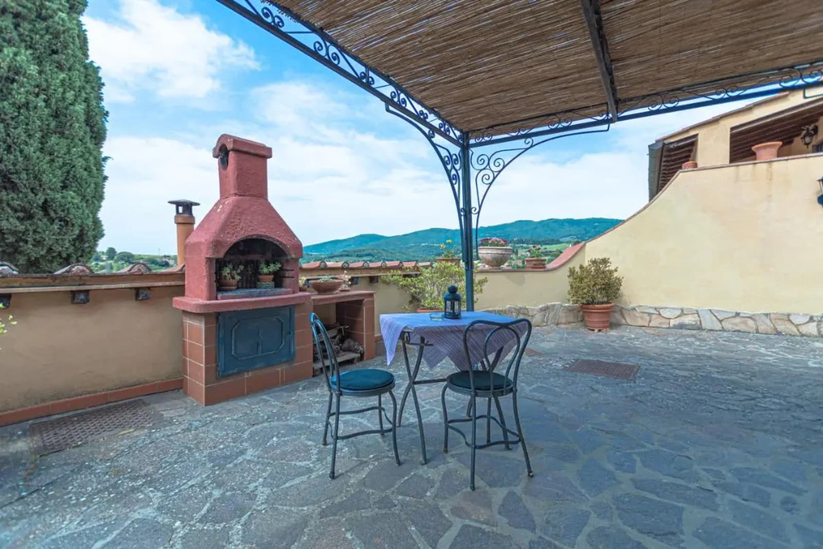 A panoramic outdoor terrace with rustic stone paving featuring a large brick barbecue fireplace in a terracotta-red finish. A small patio table covered with a light blue checkered tablecloth and two dark wrought-iron chairs with black cushions sit under a bamboo reed pergola roof supported by an ornate iron frame. The background opens to a scenic view of the green Chianti hills under a partly cloudy sky, with potted plants and traditional Tuscan villa walls framing the relaxation area.