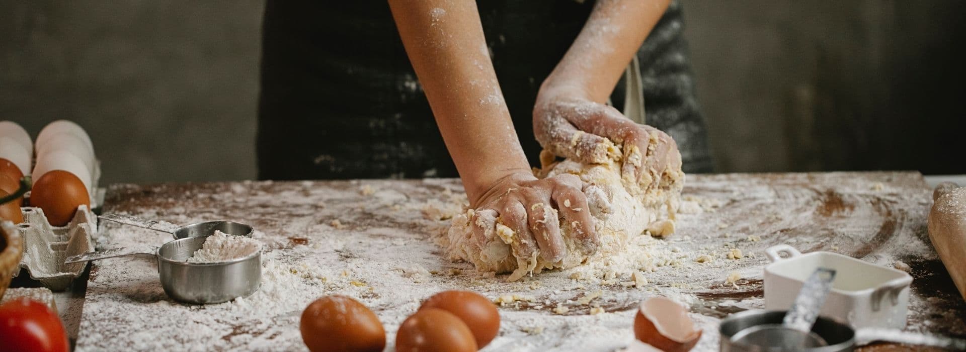 Mani infarinate che impastano la pasta fresca su un tavolo di legno cosparso di farina, con uova, gusci e un misurino in primo piano durante una cooking class in Maremma