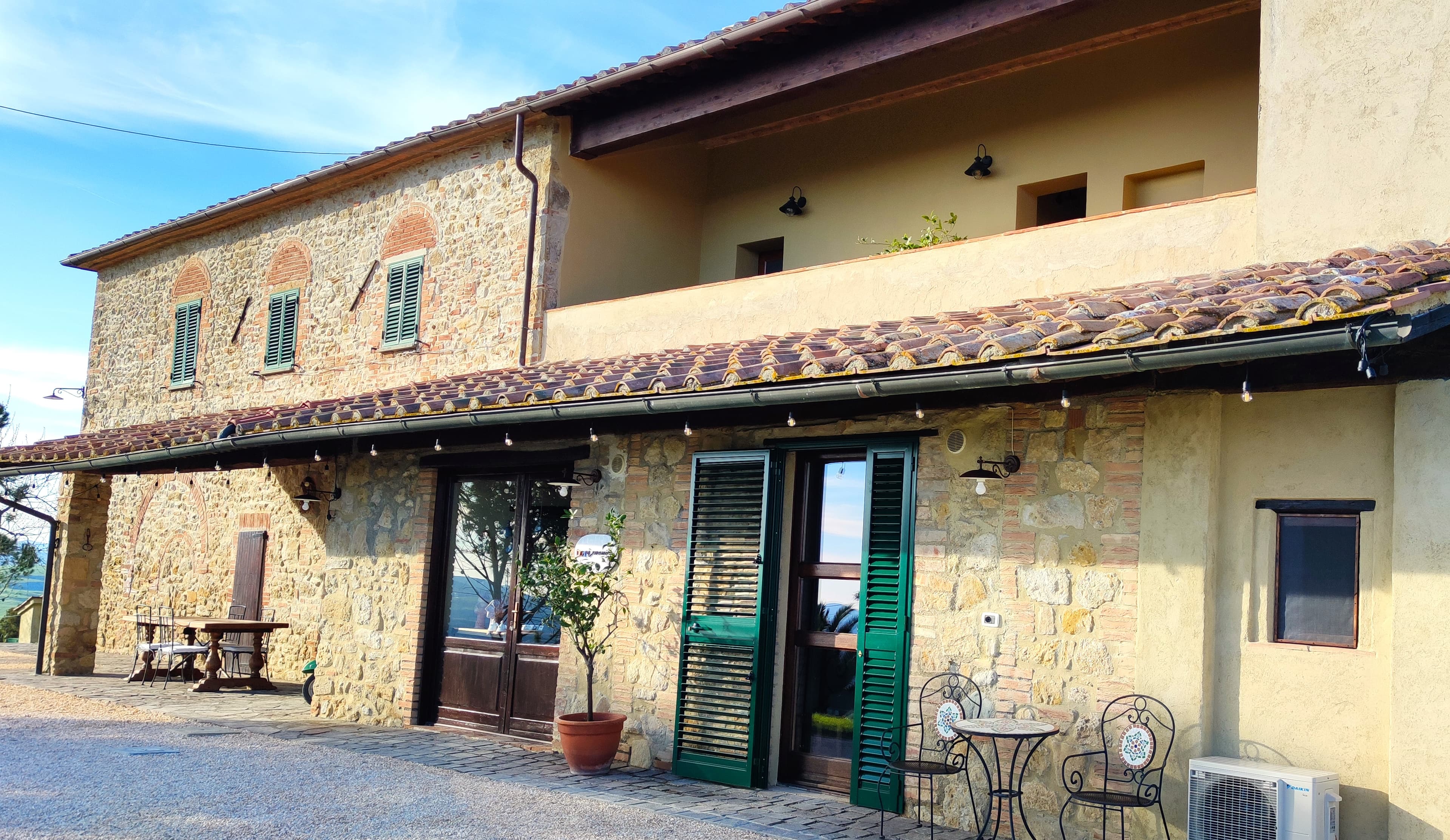 The facade of a two-story Tuscan rural house, characterized by an exposed stone section on the left with brick arches and an ochre plastered section on the right with an upper loggia. On the ground floor, a long porch with a tiled roof and wooden beams shelters a stone-paved area featuring a wooden dining table, a small tree in a terracotta pot, and a wrought-iron coffee table with two chairs. Large French doors with dark green shutters open onto the outdoors, which features a light gravel courtyard and an air conditioning unit in the right corner, all under a partly cloudy sky.