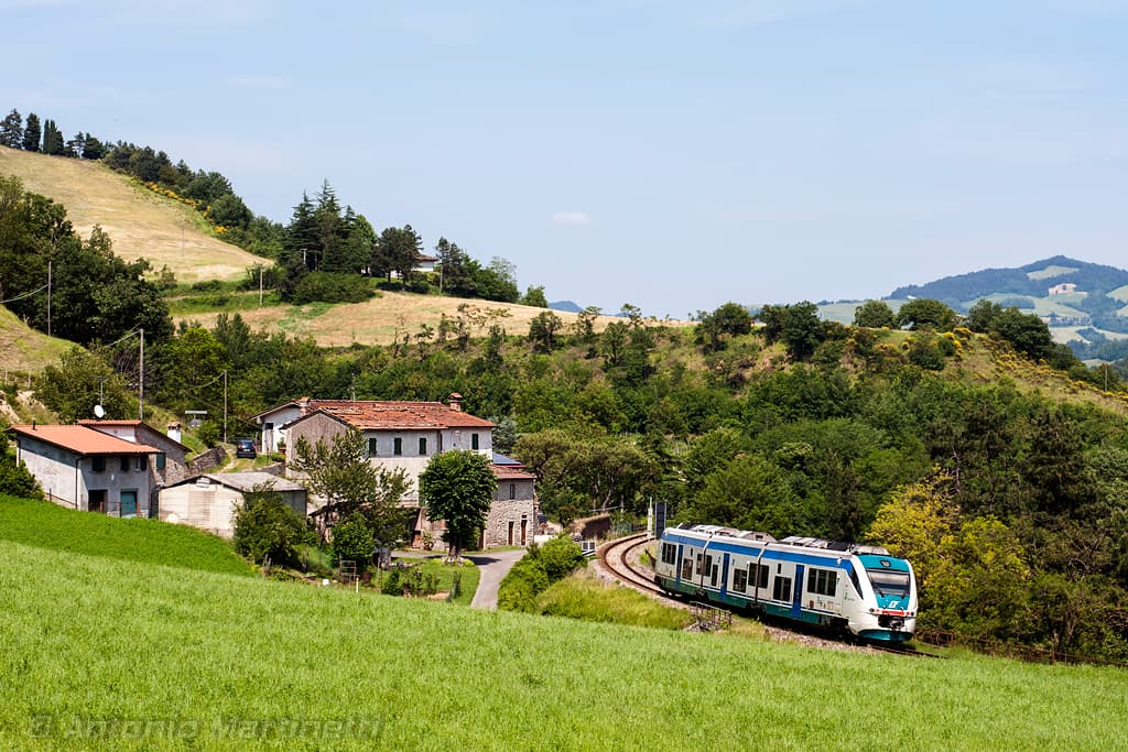 A regional train passes through the Marradi area along the Faentina line, traversing a hilly landscape with rural houses and green fields
