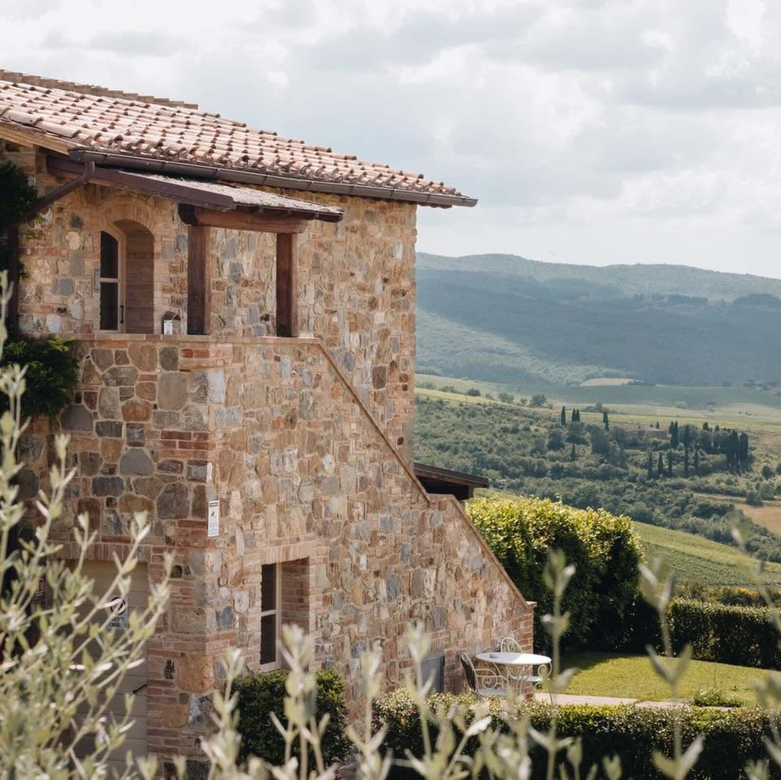 Primo piano di un tradizionale casale toscano in pietra a vista con tetto in tegole di terracotta e scale esterne. Sul balcone superiore si notano dettagli in legno rustico e una finestra ad arco. In basso, un giardino curato ospita un tavolino con sedie in ferro battuto bianco, circondato da siepi. Sullo sfondo si estende uno spettacolare panorama della campagna toscana, con colline verdi, vigneti e i classici cipressi sotto un cielo parzialmente nuvoloso.