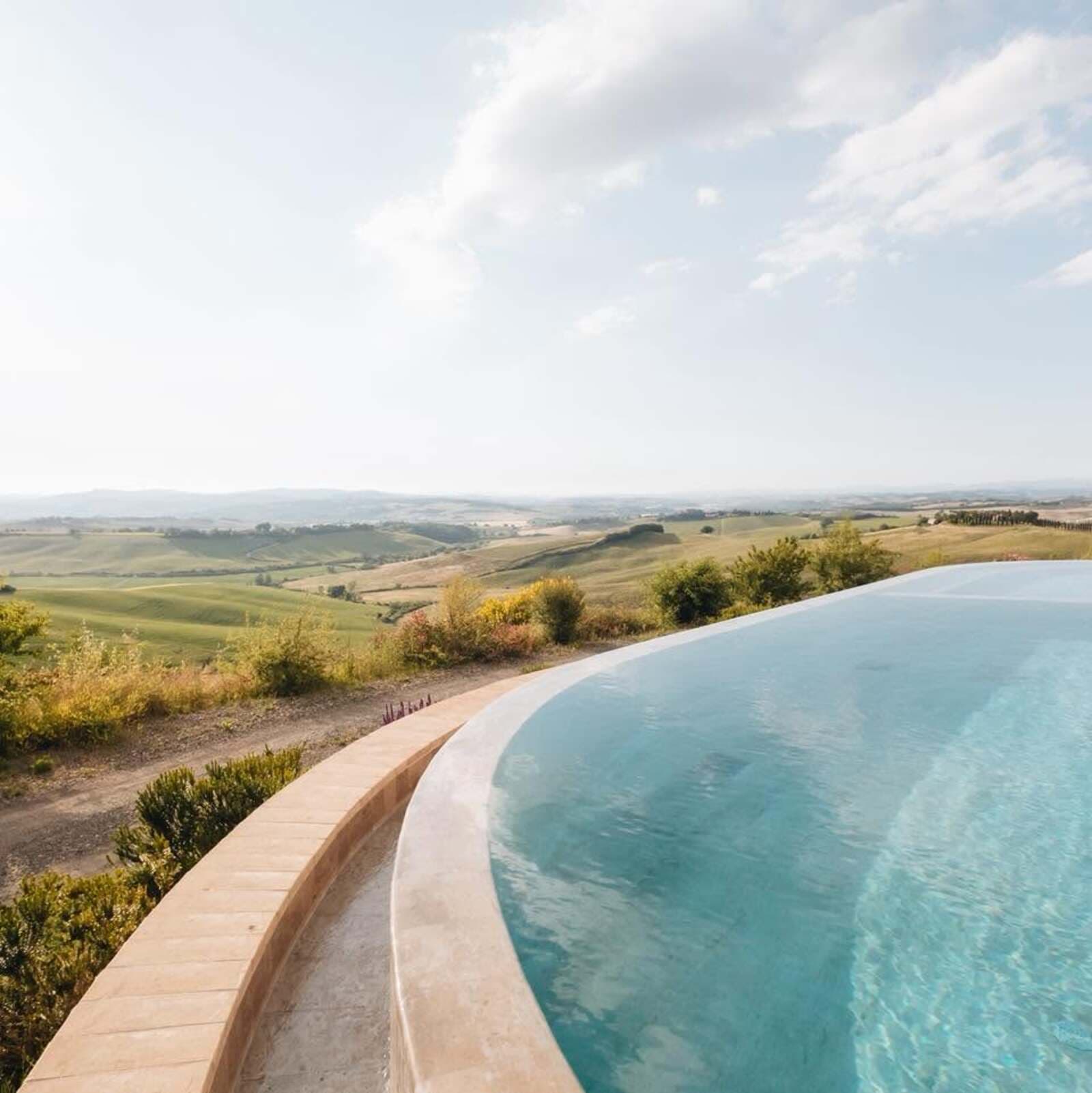 Vista ravvicinata e prospettica del bordo curvo di una piscina a sfioro con acqua cristallina e turchese. Il bordo in pietra chiara della piscina si affaccia direttamente su uno spettacolare panorama collinare toscano, caratterizzato da ampie distese di campi verdi e dorati, tipici della Val d'Orcia o delle Crete Senesi. Sullo sfondo si estendono colline ondulate a perdita d'occhio sotto un cielo azzurro e luminoso, punteggiato da nuvole bianche e leggere. La vegetazione spontanea e piccoli arbusti mediterranei costeggiano il perimetro esterno della struttura.