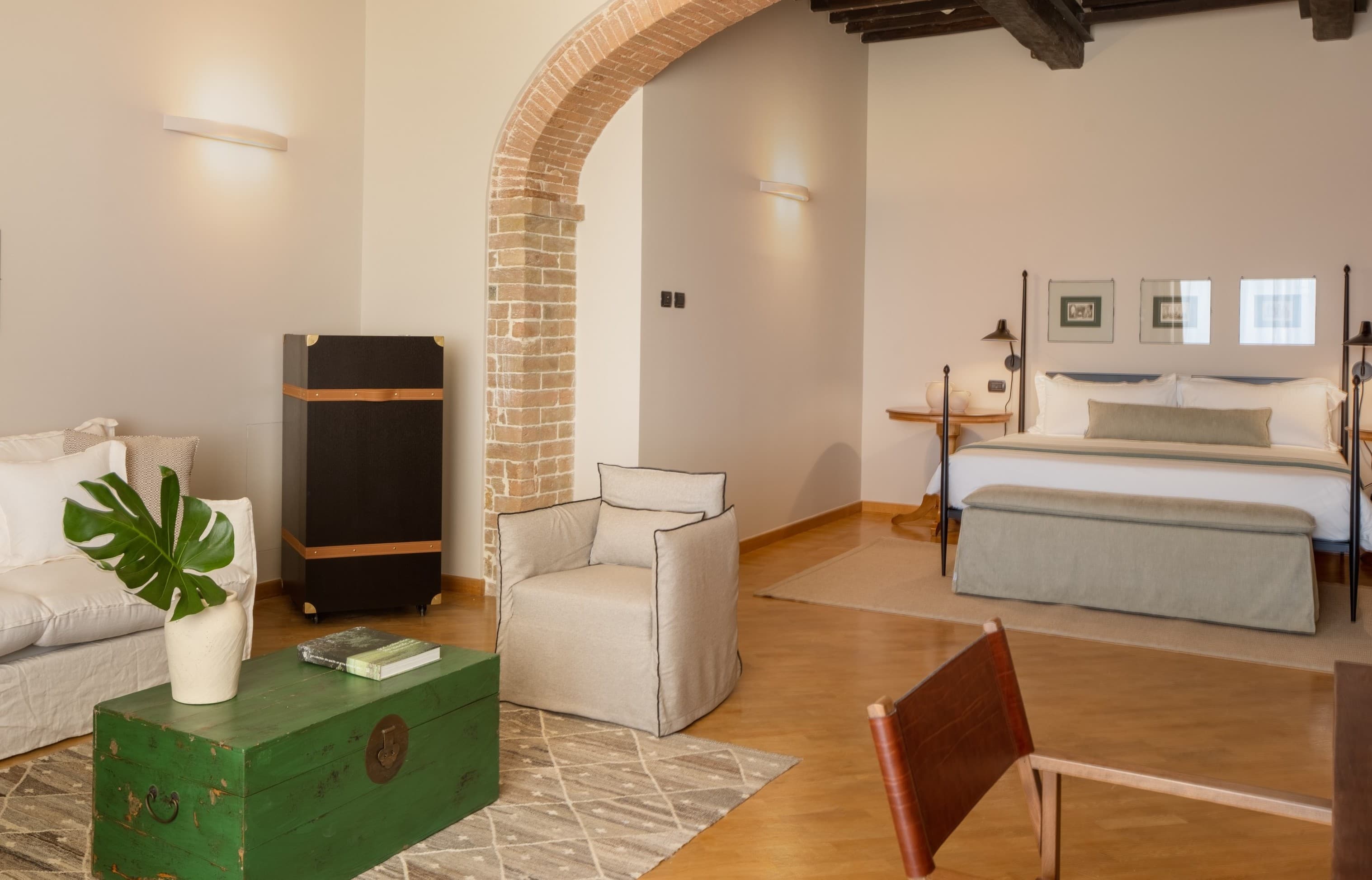 A perspective view of a spacious and luxurious open-plan Tuscan suite. In the foreground, a sitting area featuring a white linen sofa, a light grey upholstered armchair and an antique green trunk-style coffee table with a Monstera plant and an art book. The sitting area is separated from the sleeping area by a majestic exposed brick arch. In the background stands a modern black wrought-iron four-poster bed with immaculate white bed linen and a footboard bench. Above the headboard, three framed botanical prints are perfectly aligned. The round wooden bedside tables are fitted with elegant table lamps. The warm lighting from the wall sconces and bedside lamps creates a cosy and sophisticated atmosphere, highlighting the exposed dark wooden beams on the ceiling and the parquet flooring.  Translated with DeepL.com (free version)