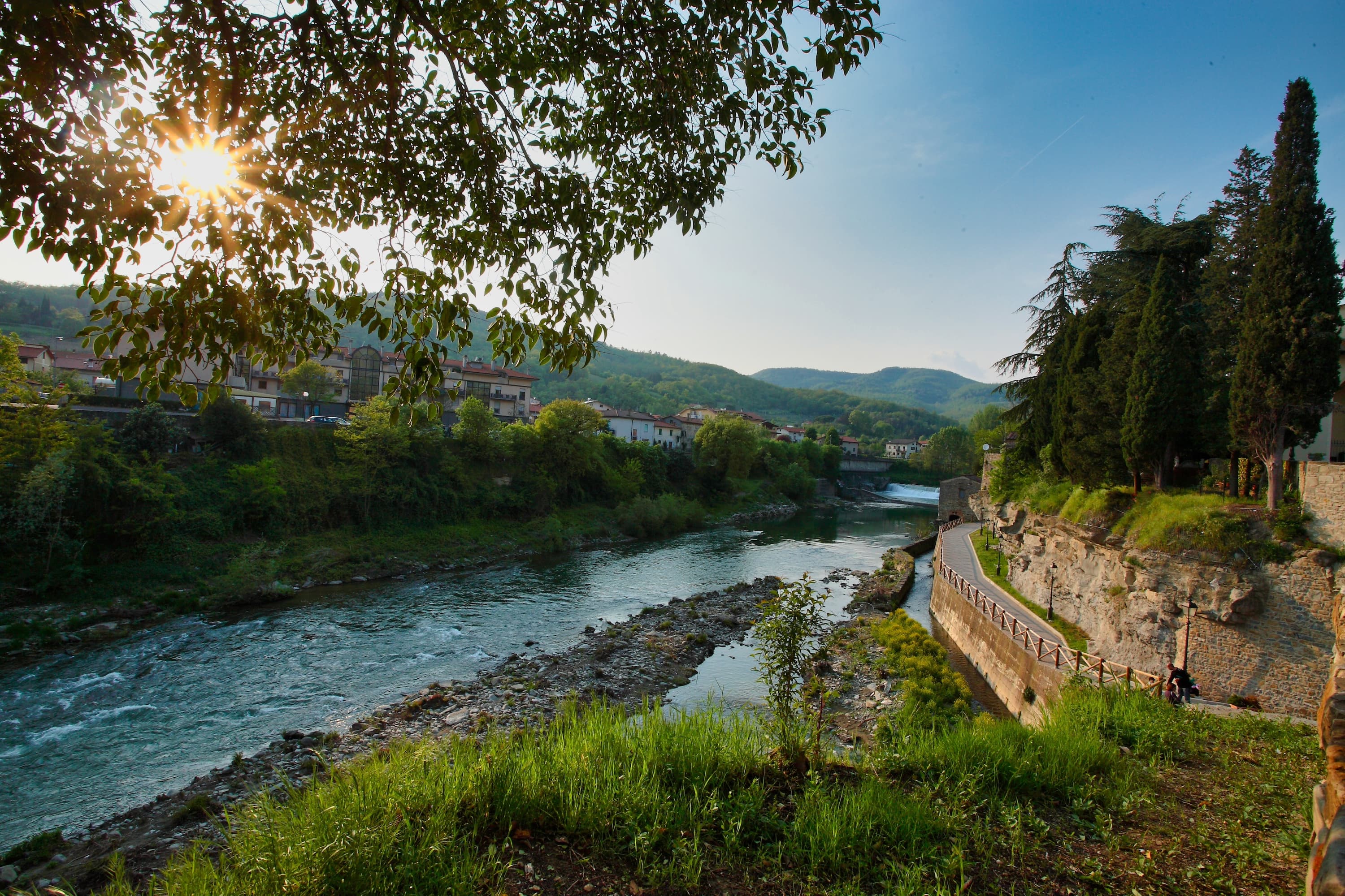 Panorama al tramonto sul fiume Arno a Subbiano nel Casentino: acque turchesi tra rive verdi, un percorso pedonale e ciclabile affiancato da un muro in pietra, colline boscose e un borgo sullo sfondo illuminato dalla luce radente del sole