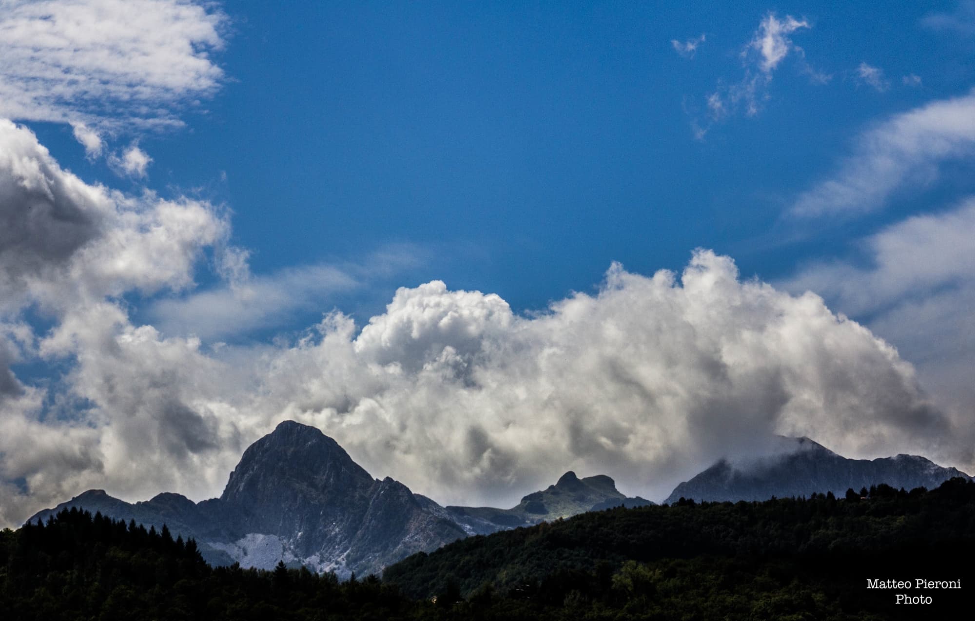 Panorama delle Alpi Apuane con le cime di Omo Morto e Pania, montagne rocciose che si stagliano contro il cielo, circondate da vegetazione e vallate.