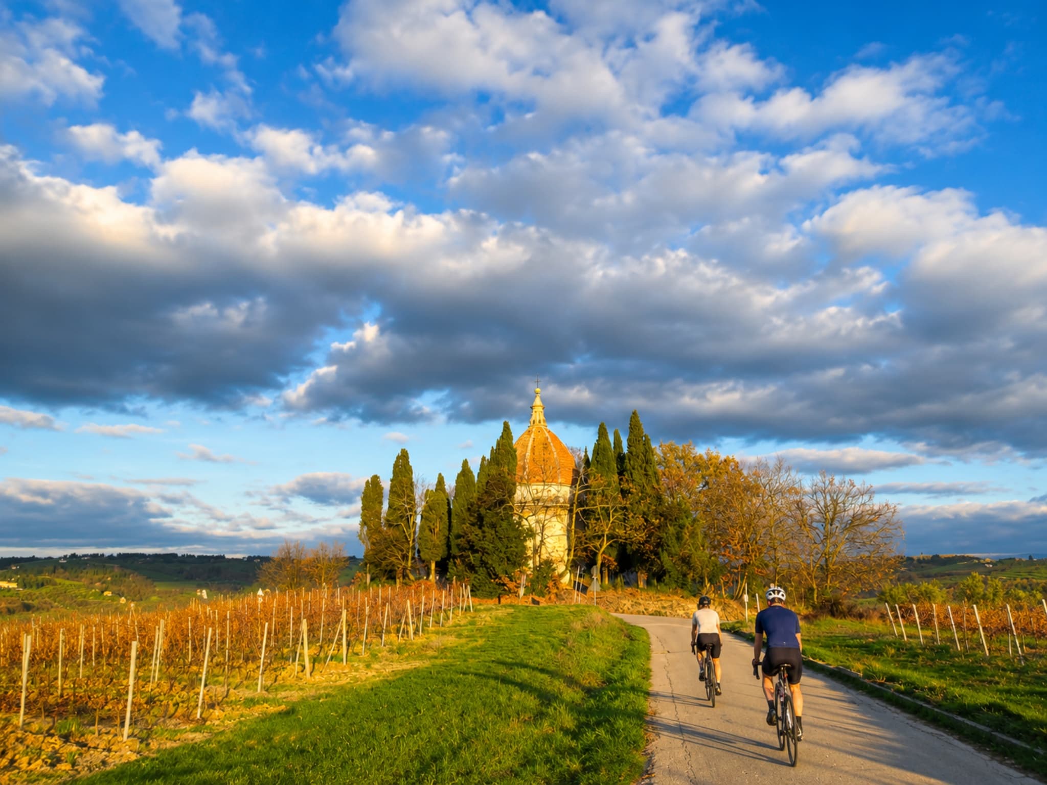 Two cyclists on e-bikes approach the Chapel of San Michele Arcangelo in Semifonte, nestled among Tuscan vineyards at sunset