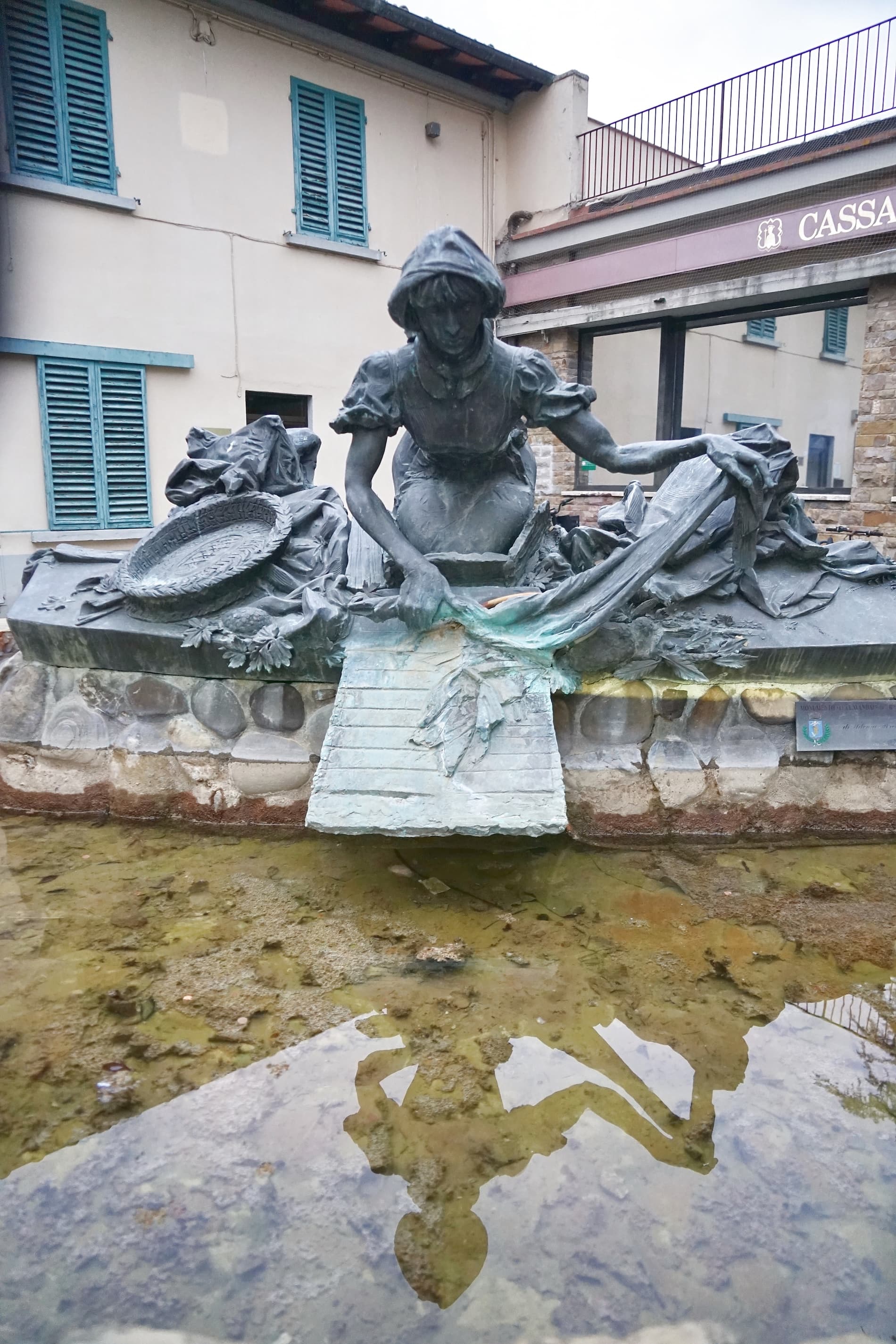 Estatua de bronce del monumento a la Lavandera en Grassina, que representa a una mujer arrodillada lavando la ropa en una tabla de madera colocada sobre una pila de agua.