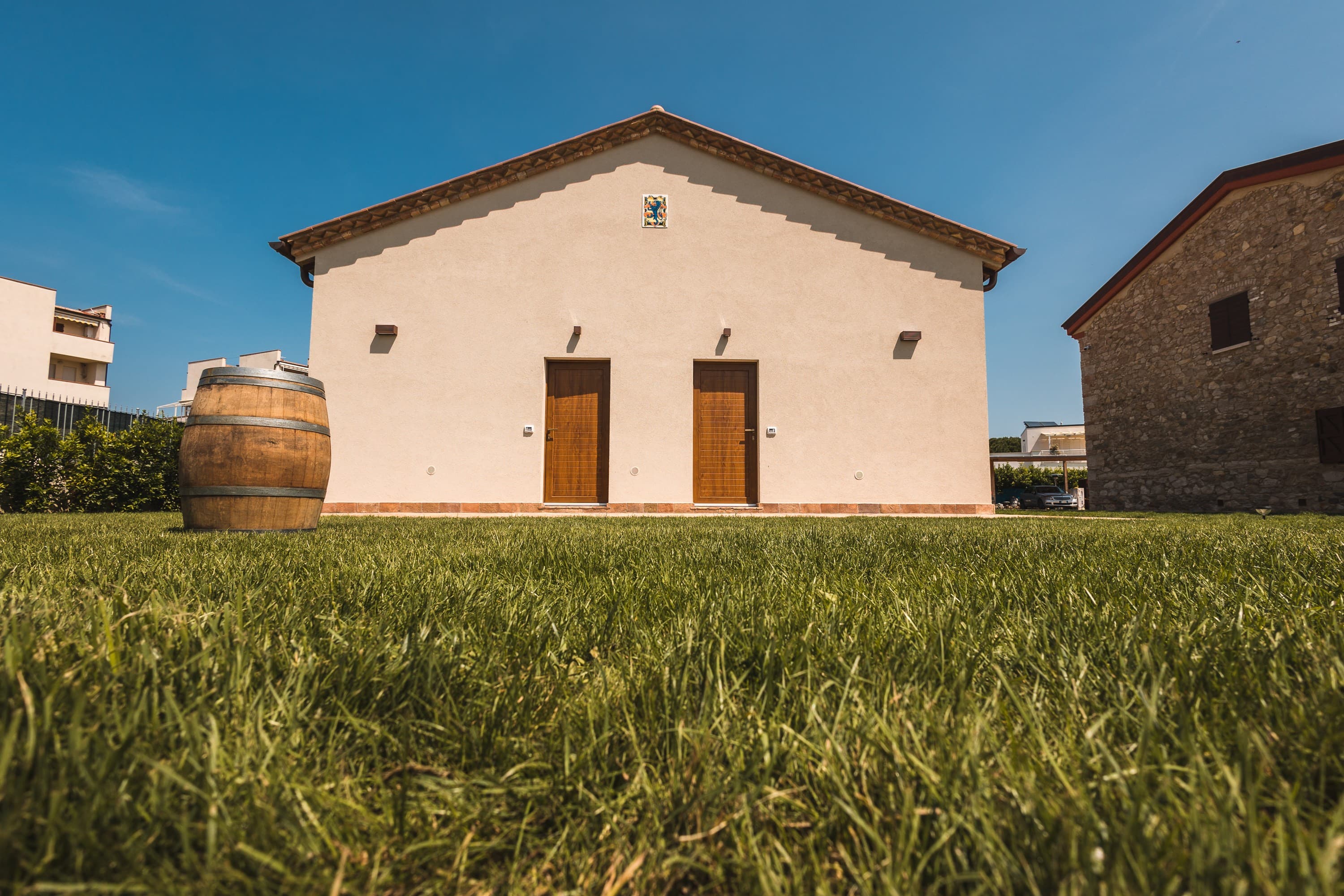 Low-angle view of a well-manicured green lawn surrounding a Tuscan-style property or winery. In the background stands the symmetrical facade of a light-colored building, featuring two separate dark wood entrance doors and a gabled roof. A large, decorative wooden wine barrel sits on the left side of the grass, while the stone wall of an adjacent building is visible on the right, all under a clear and bright summer sky.