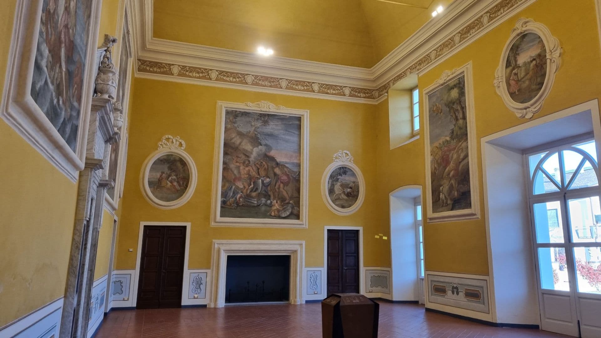 Salone affrescato del Castello di Pallerone con pareti gialle, grandi dipinti storici, camino centrale e finestre ad arco.