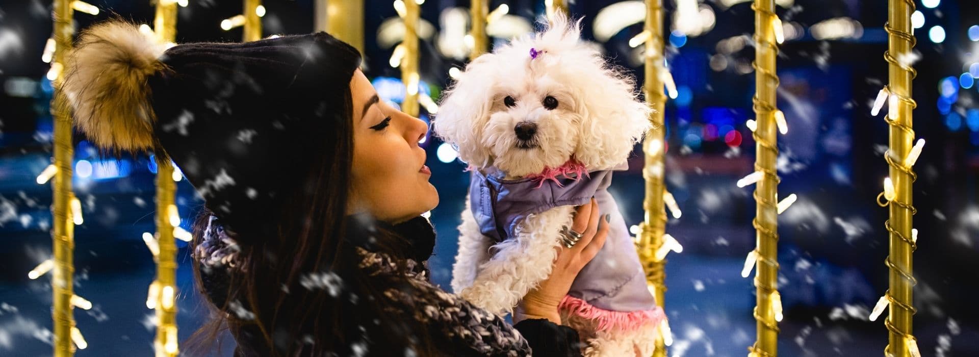 Donna con berretto invernale bordato di pelliccia che tiene tra le braccia un cane maltese vestito con un cappottino, tra luci dorate scintillanti e neve che cade, durante una vacanza di Capodanno pet-friendly in Toscana