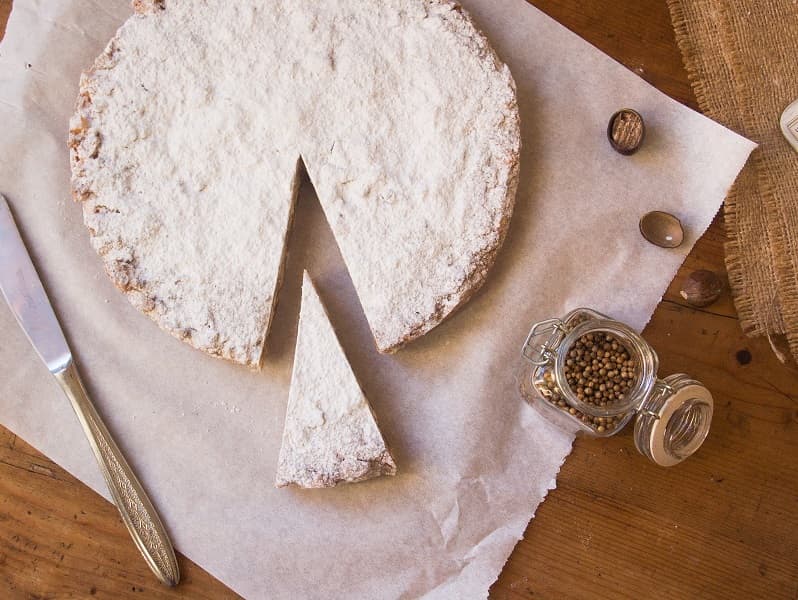 Ein mit Puderzucker bestreuter Panforte, bei der eine Scheibe fehlt. Die für die Zubereitung verwendeten Gewürze sind auch auf dem Tisch zu sehen.
