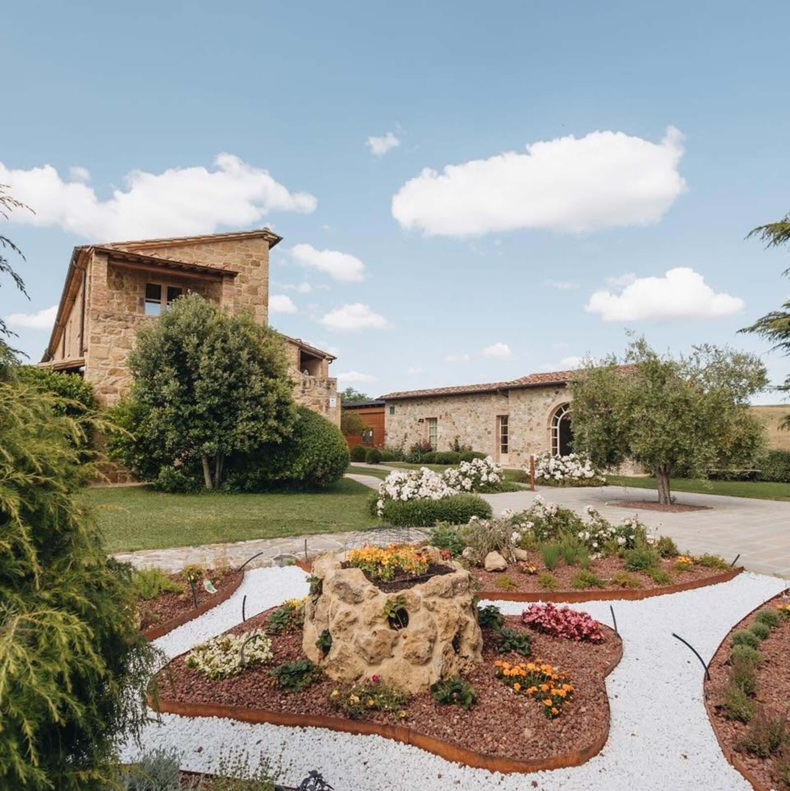 Veduta panoramica del giardino curato di un borgo o agriturismo in Toscana, caratterizzato da tradizionali casali in pietra a vista con tetti in tegole. In primo piano spiccano delle aiuole fiorite geometriche delimitate da bordure metalliche, riempite di pacciamatura scura e circondate da vialetti in ghiaia bianca. Al centro si trova un antico pozzo in pietra decorato con fiori. Sullo sfondo si sviluppa un grande prato verde con alberi di olivo, cespugli di rose bianche e i camminamenti pavimentati che collegano le strutture ricettive sotto un cielo azzurro con nuvole bianche.