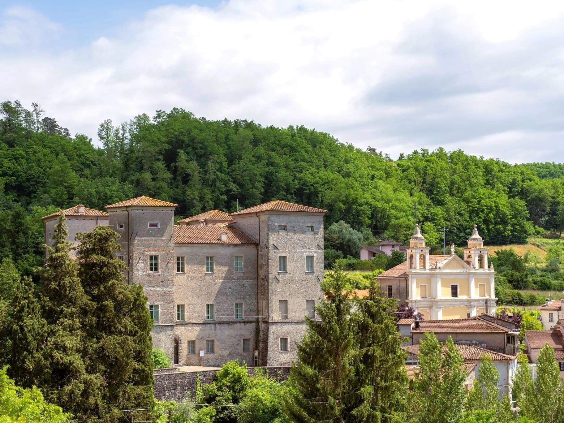 Vista panoramica del borgo di Pontebosio con il Castello dei Malaspina in primo piano, la chiesa barocca gialla con campanile e il bosco verde sullo sfondo, nella Valle del Taverone in Lunigiana