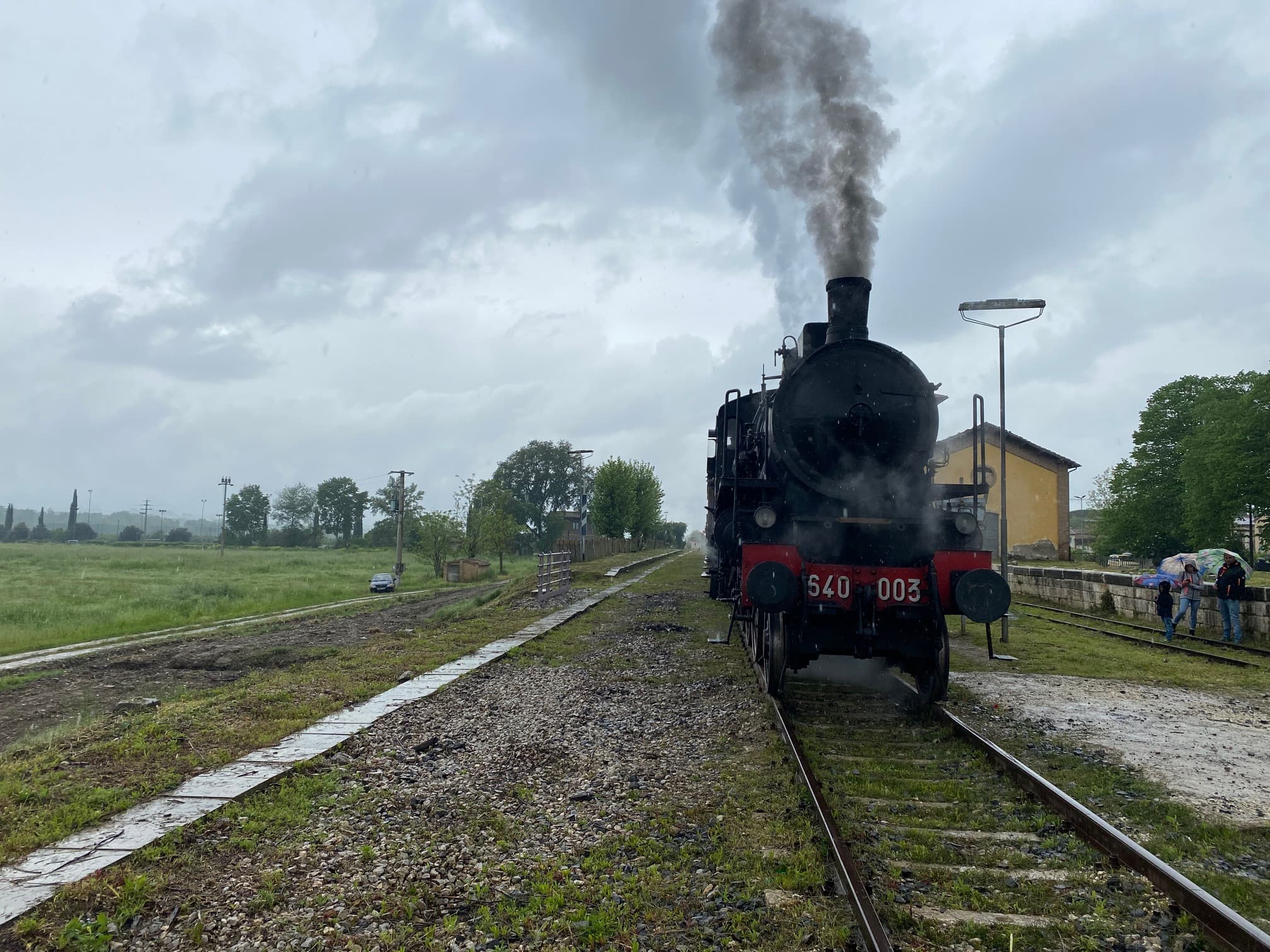 Locomotiva a vapore 640-003 del Treno Natura ferma sui binari di una piccola stazione toscana, con il fumo nero che sale dalla ciminiera e il cielo nuvoloso sullo sfondo, durante l'escursione storica da Siena ad Abbadia San Salvatore