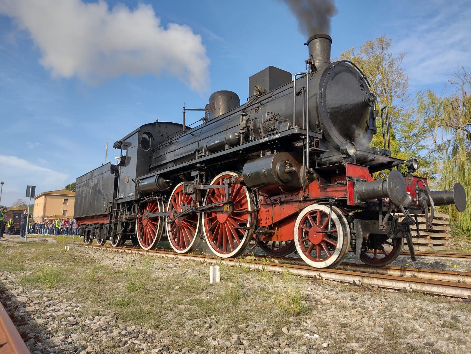 Locomotiva a vapore nera con ruote rosse del Treno Natura ferma sui binari, con pennacchio di fumo bianco e visitatori sullo sfondo, durante l'escursione storica da Siena a San Giovanni d'Asso