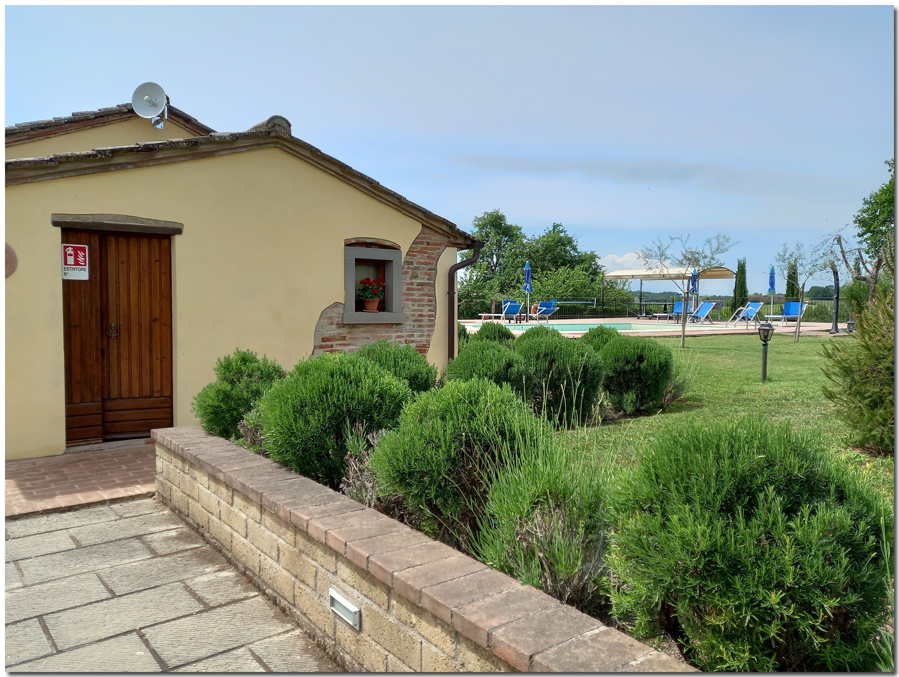 Exterior view of a Tuscan cottage outbuilding with yellow stucco walls, exposed brick details, and a dark wooden door. In the foreground, a low stone retaining wall borders a garden bed filled with lush, rounded green rosemary and lavender bushes. In the background, the spacious green lawn leads to the outdoor sun deck area, equipped with blue sun loungers, umbrellas, and a gazebo next to the private swimming pool.