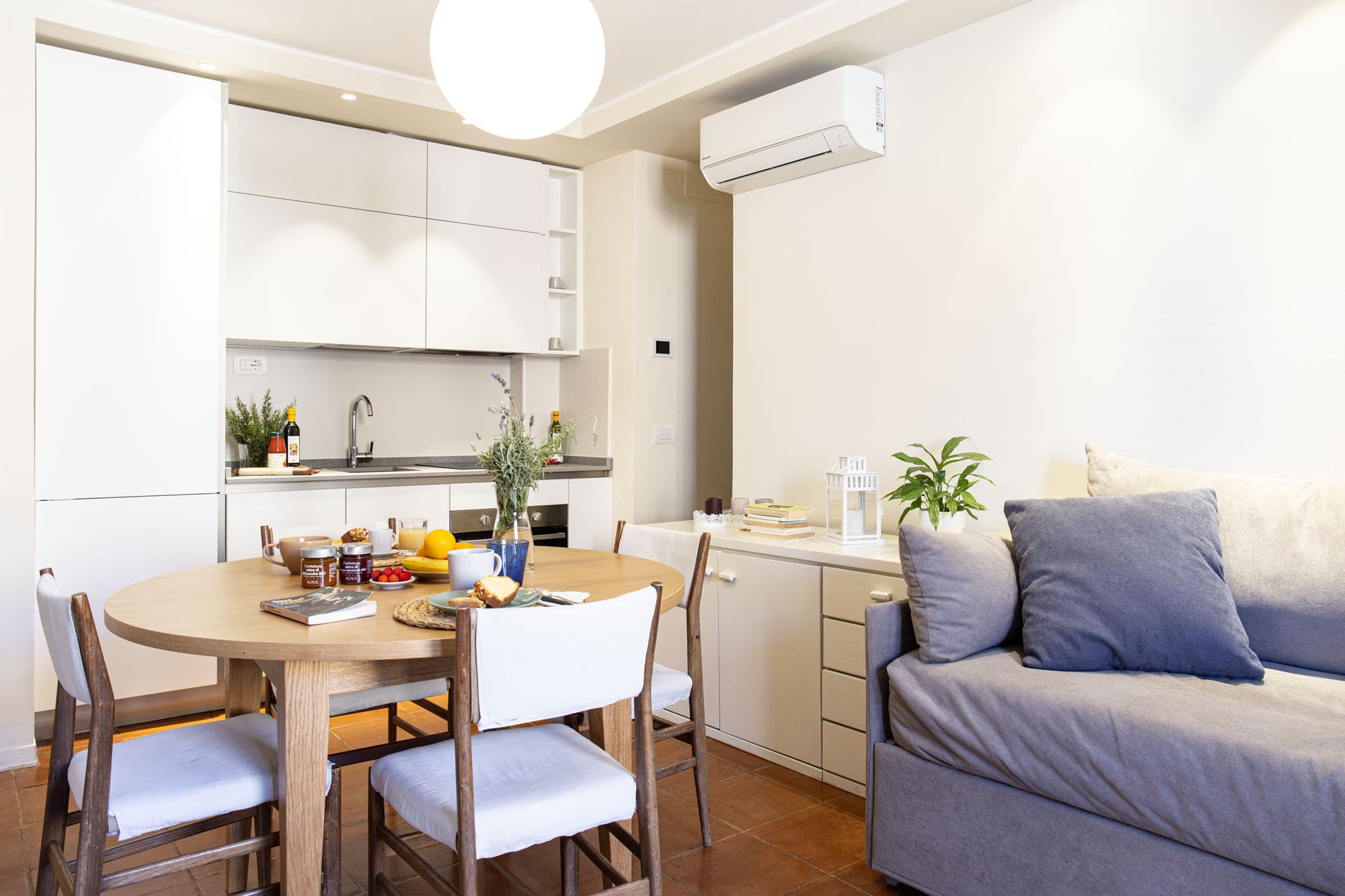 Bright interior view of an open-plan living and dining area in a holiday rental. In the foreground, a round wooden dining table is set for breakfast with mugs, fresh fruit, and jams, surrounded by four chairs with light cushions. On the right, a grey sofa bed with blue pillows stands next to a white sideboard decorated with a plant and books. In the background, a modern and linear white fully equipped kitchenette is visible underneath a wall-mounted air conditioner. The room features traditional terracotta tiled flooring.