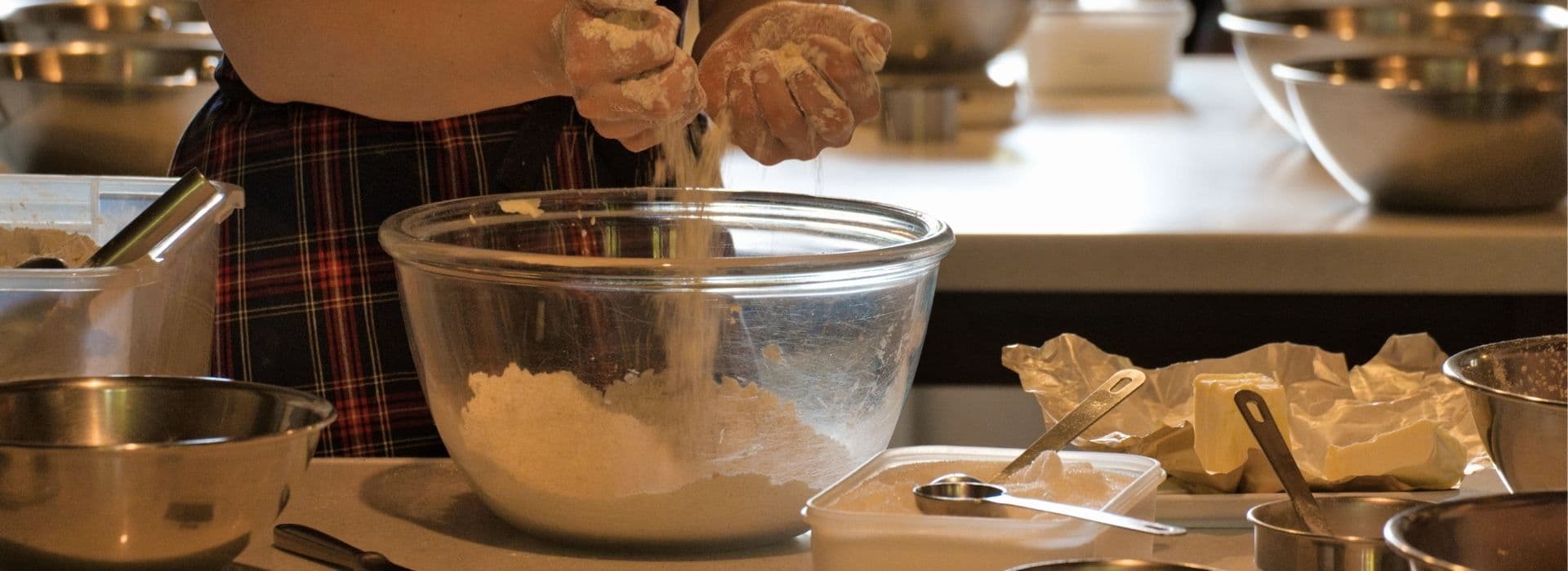 Mani infarinate setacciano farina in una ciotola di vetro durante una cooking class in Maremma, con burro, utensili e ciotole in acciaio sul piano di lavoro