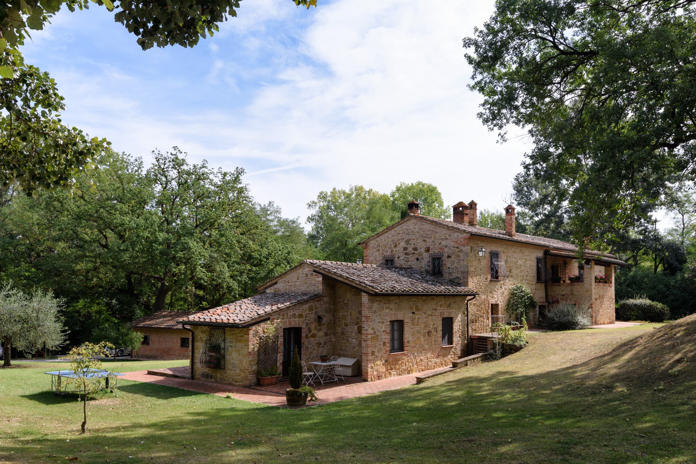 Panoramic view of the beautifully restored historic stone mill, surrounded by a large green lawn and mature trees. The property features a terracotta patio with outdoor seating, alongside a table tennis setup on the grass under a bright, partly cloudy sky.