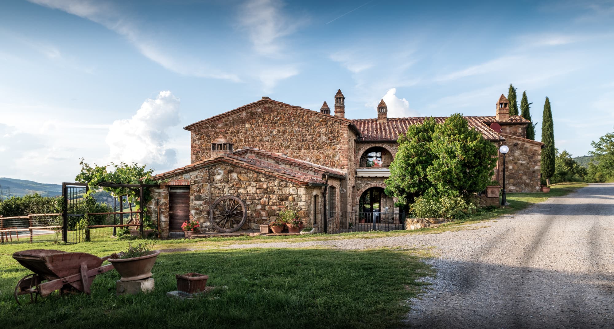 Antico casale in pietra toscano con cortile in erba, vialetto in ghiaia, una vecchia carriola in legno decorativa e vasi di fiori.
