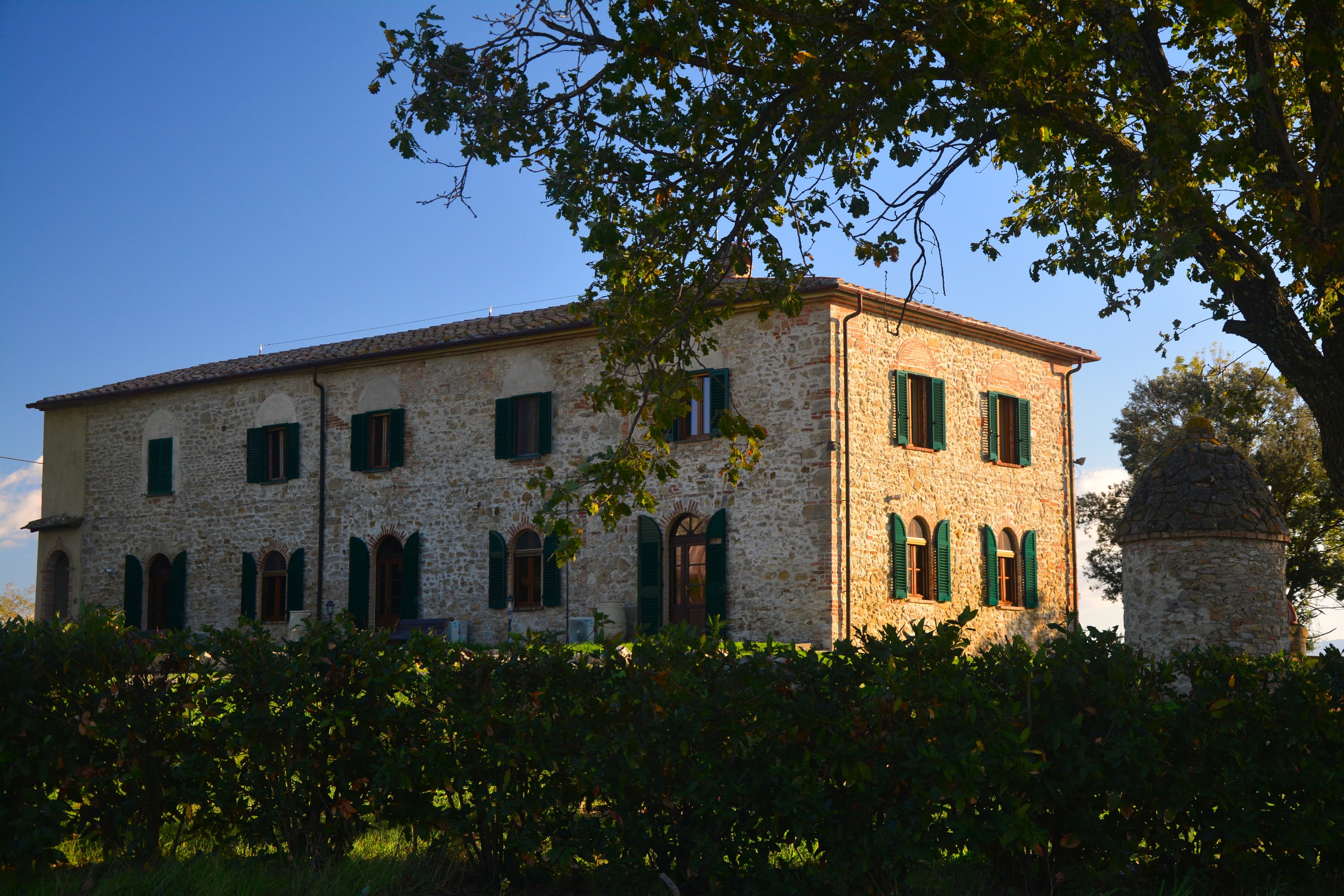 casa colonica toscana in pietra a vista a due piani, illuminata dalla luce calda del sole. L'edificio presenta numerose finestre e porte centinate al piano terra, tutte dotate di persiane in legno verde scuro.  La proprietà è parzialmente nascosta in primo piano da una fitta siepe verde e incorniciata dalle fronde di un albero sulla destra, accanto al quale sorge una caratteristica torretta o antico pozzo in pietra con cupola conica. Sullo sfondo si staglia un cielo azzurro e limpido