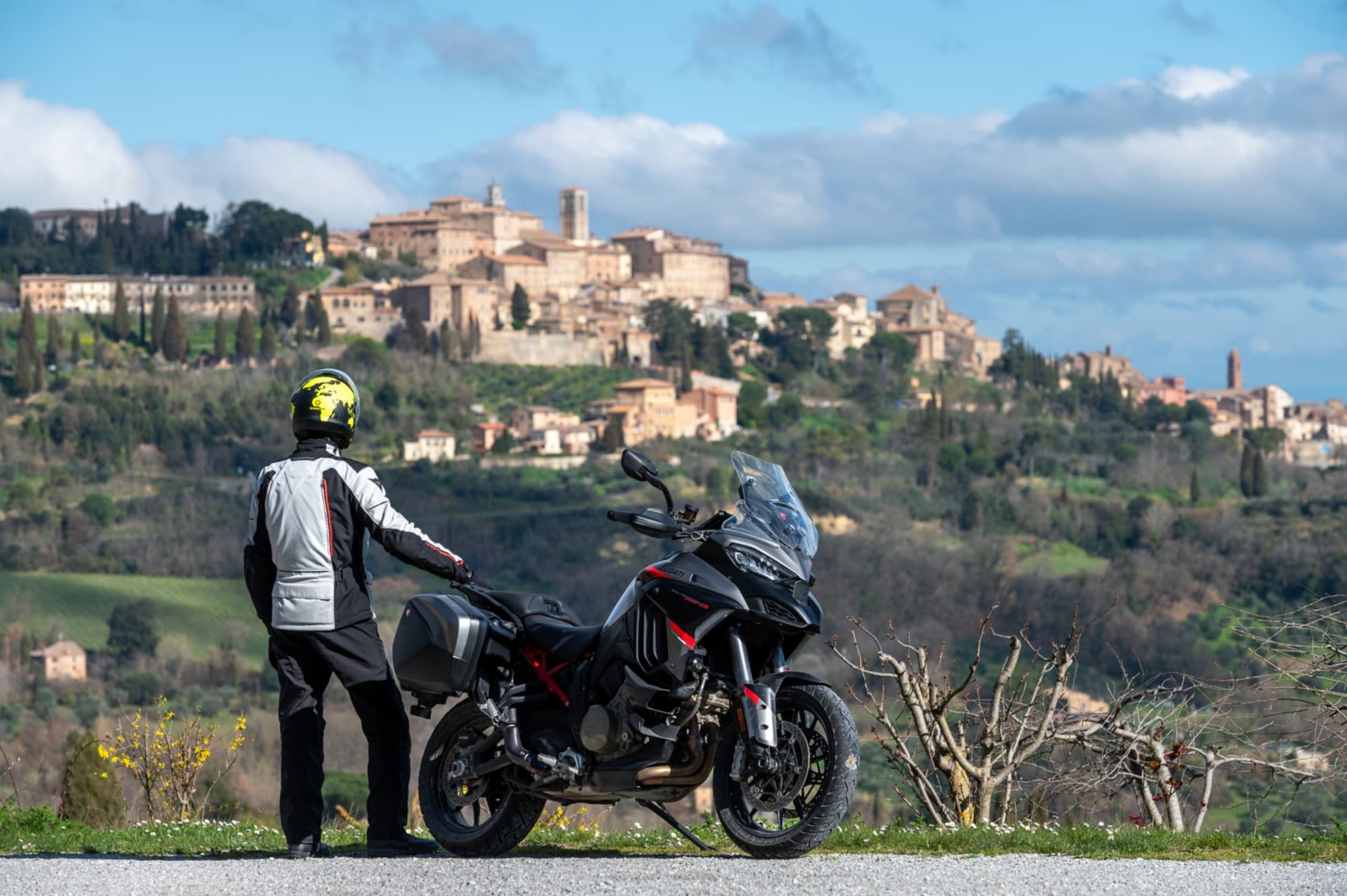 A motorcyclist admiring the scenery from the side of their motorcycle, with the village of Montepulciano in the background.