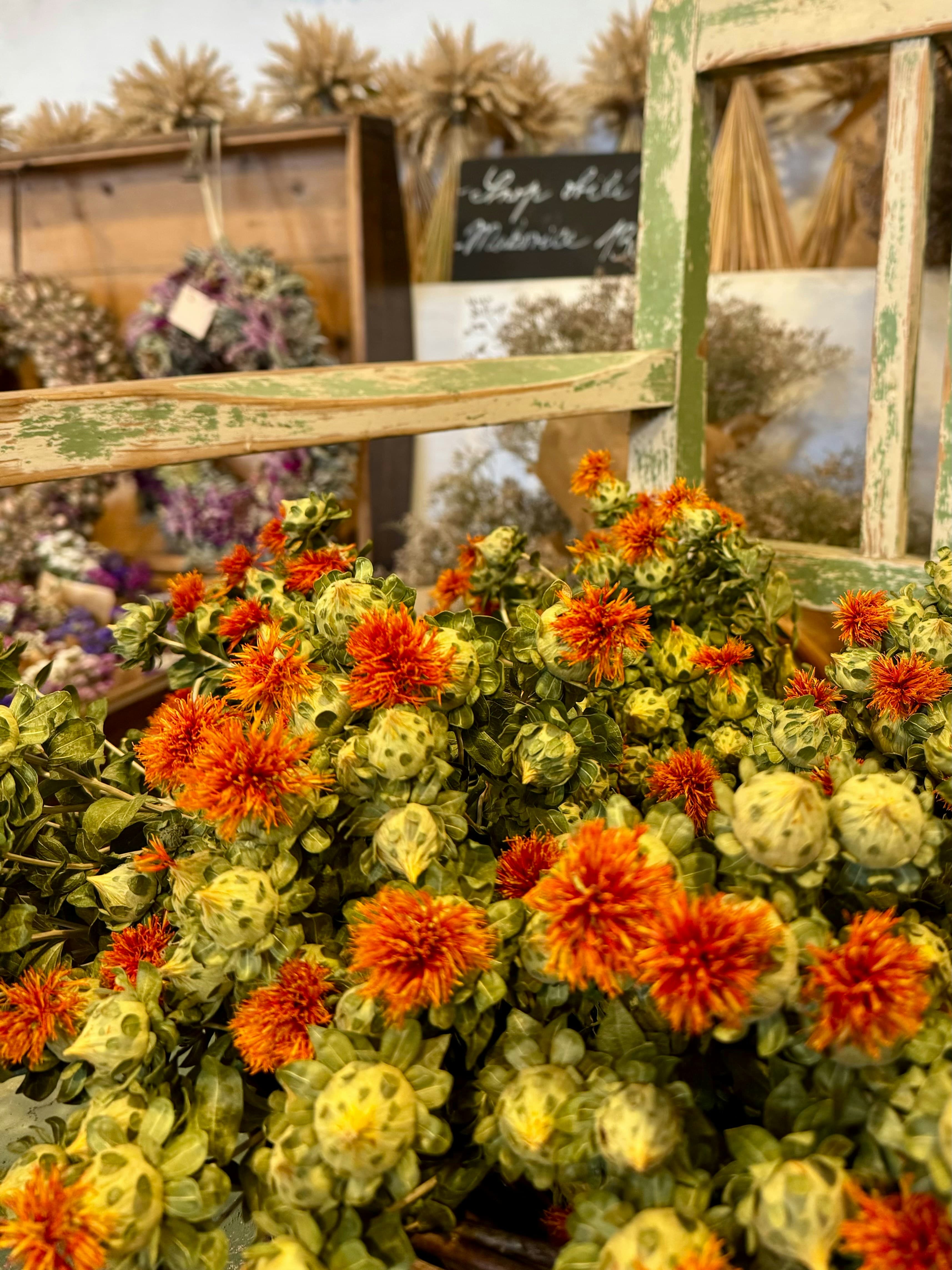 Close-up of orange flowers and green buds displayed in a vintage wooden crate inside an artisanal flower shop, with dried flower arrangements in the background