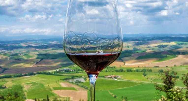 Bicchiere di Brunello di Montalcino in primo piano, con la campagna toscana sullo sfondo tra vigneti e dolci colline.