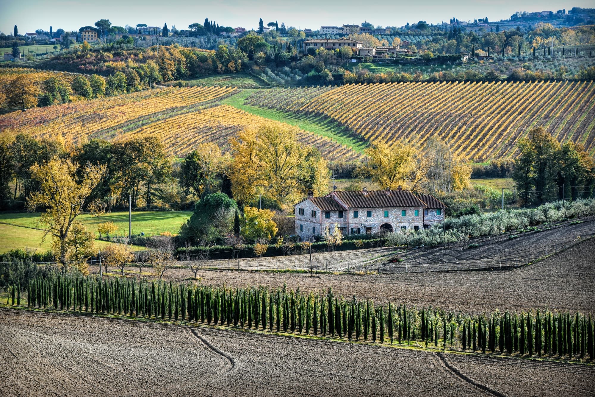 Paisaje rural en la Vía Francigena toscana: casa de labranza de piedra enclavada entre campos arados, hileras de cipreses y viñedos dorados.