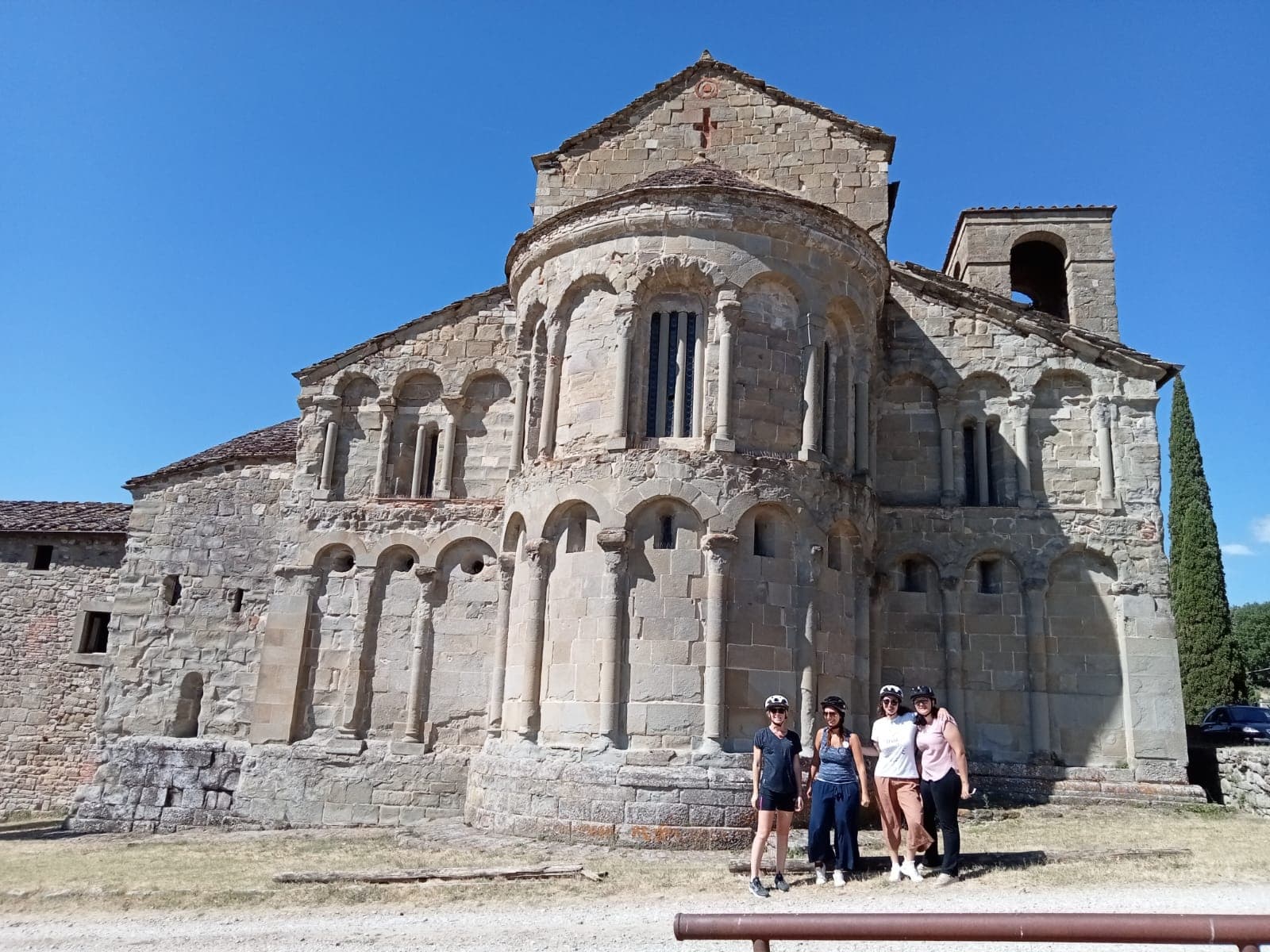 Four cyclists wearing helmets pose in front of the Romanesque apse of the Pieve di Romena, a sandstone building with blind arches and a bell tower, on a sunny day in Casentino